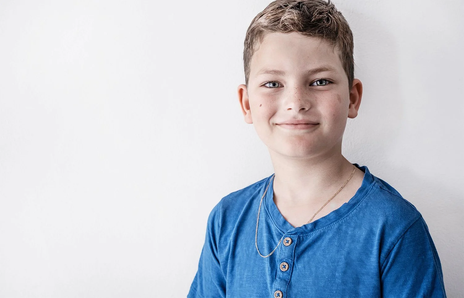 A young boy with short brown hair, blue eyes, and freckles, smiling, wearing a blue shirt with buttons and a silver chain necklace, standing against a plain white background.