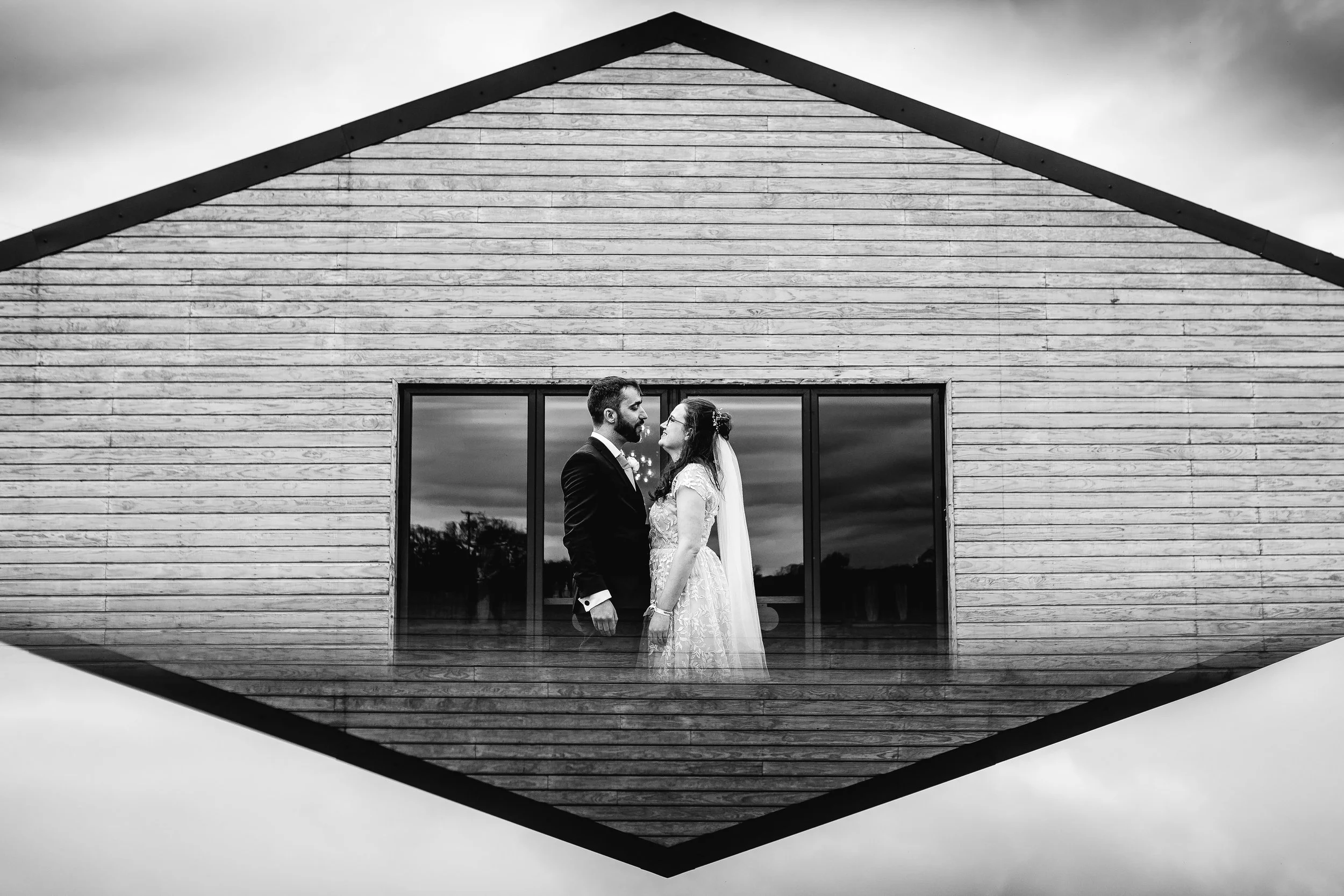 A black and white photo of a bride and groom standing inside a building with large windows, viewed through a reflective glass that creates a diamond-shaped frame around them, with cloudy sky reflections outside.
