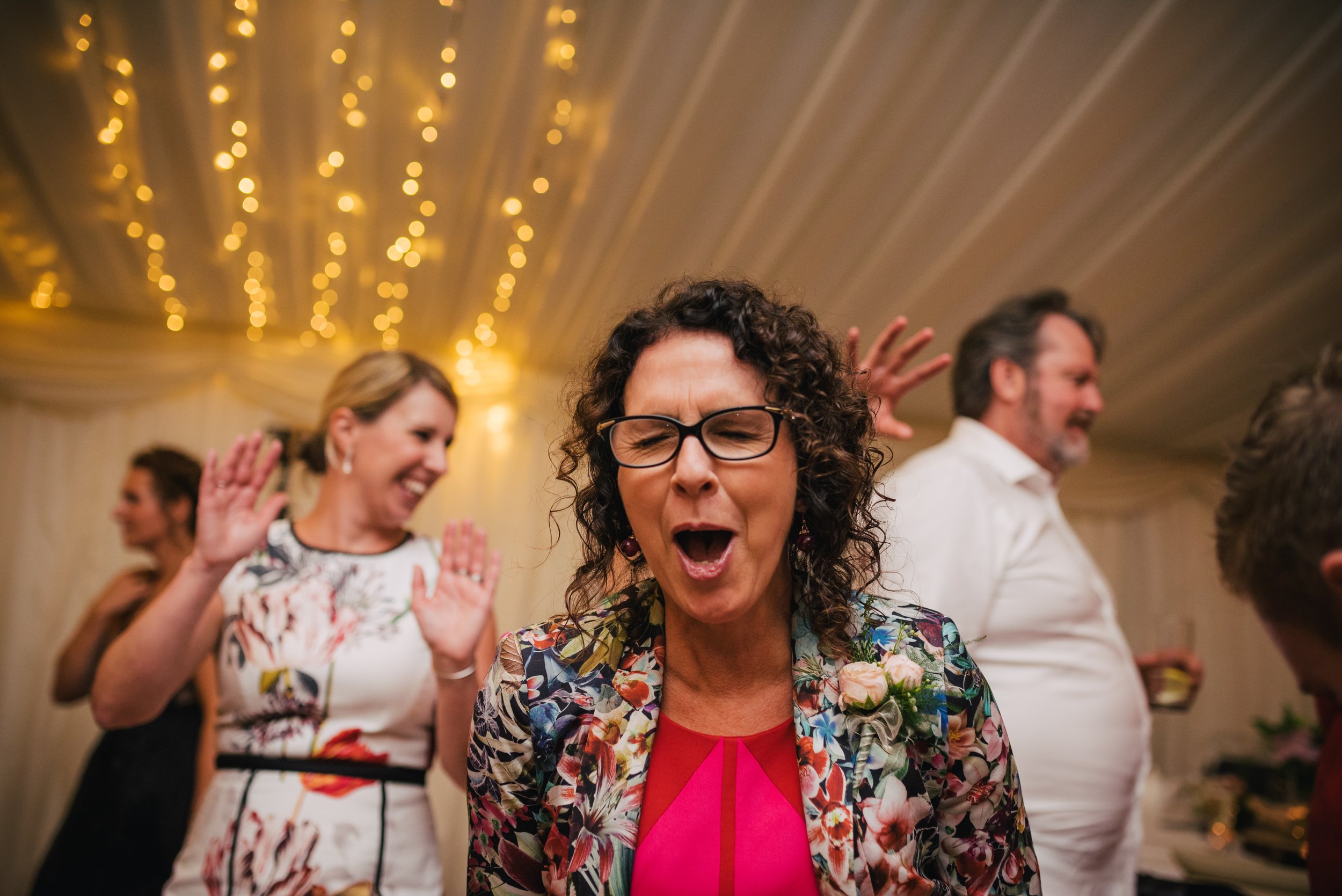 A woman with curly dark hair, glasses, and a floral blazer singing or shouting at a lively celebration, with other guests in the background under warm string lights.