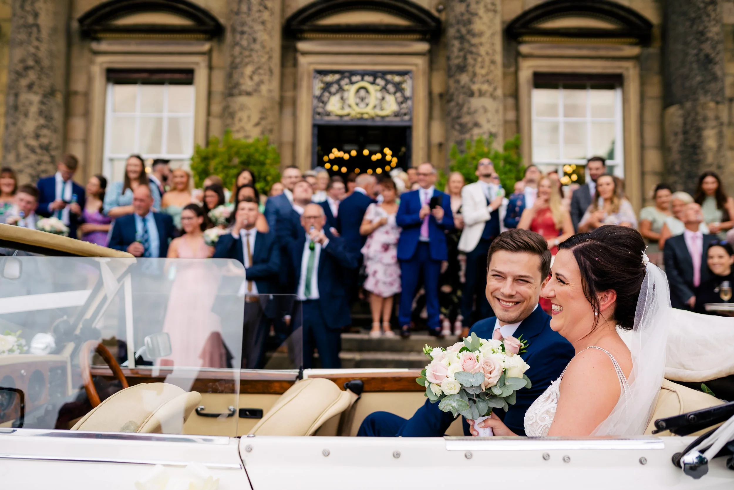 A bride and groom sitting in a vintage car, smiling, with a large wedding party and guests gathered behind them on the steps of a historic building.