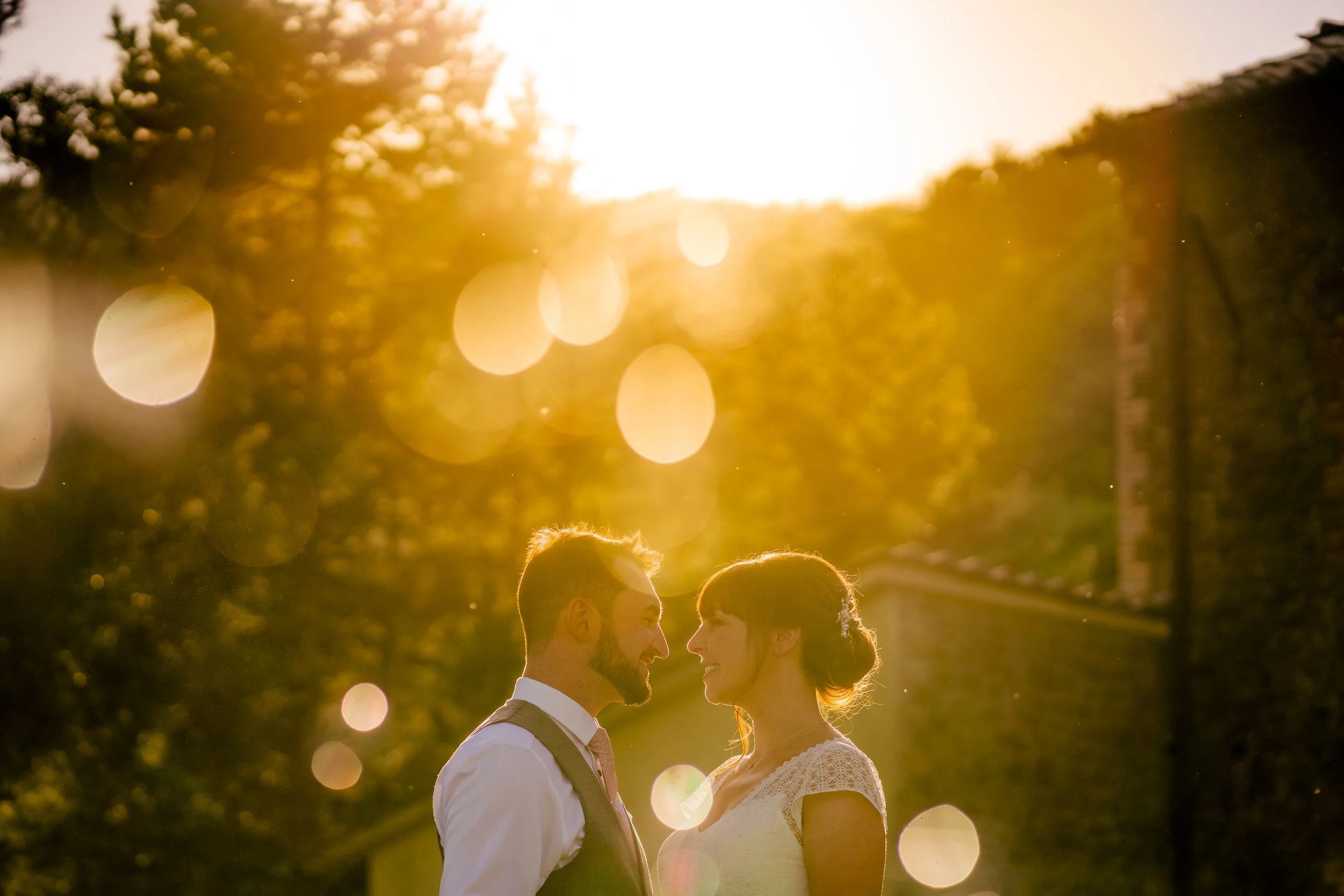 A couple smiling and looking at each other during sunset outdoors, with trees and a brick wall in the background, and light flares creating a warm glow.