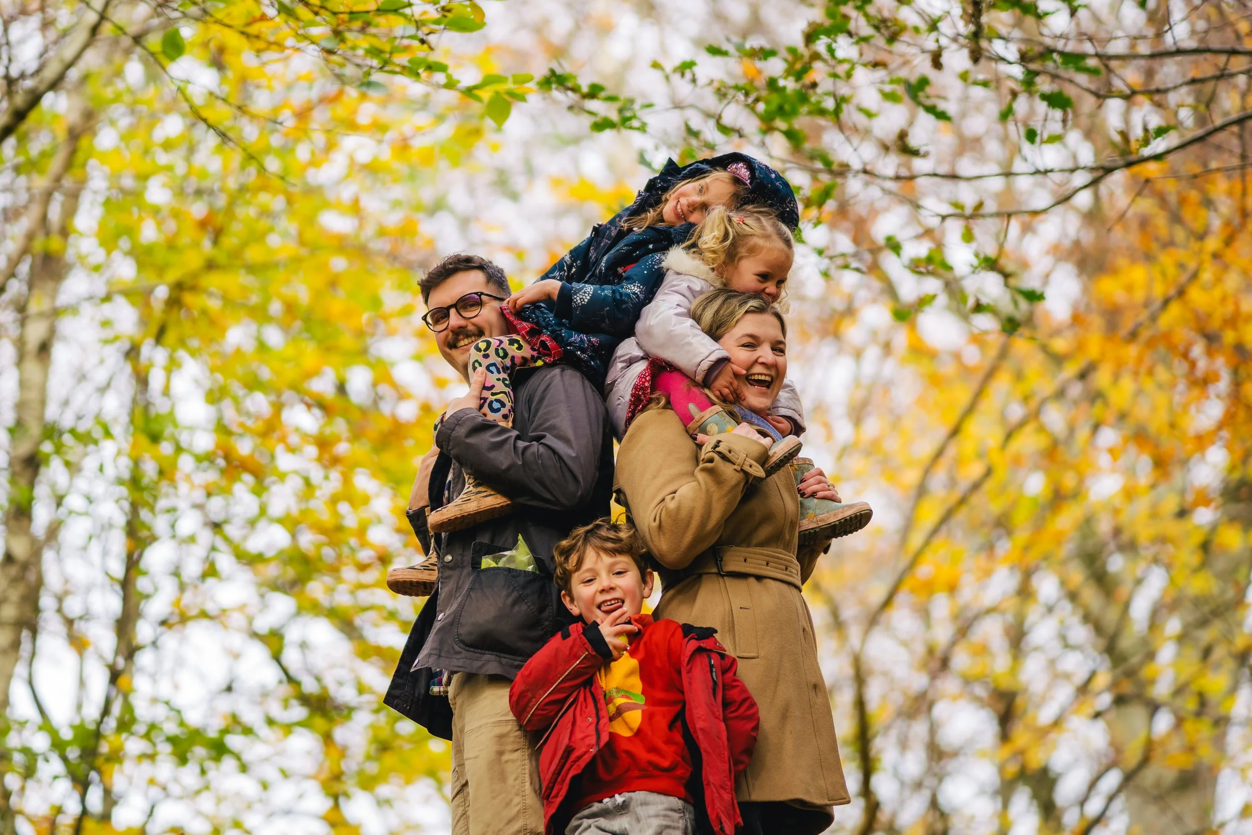 Family of five, including adults and children, on a hike in autumn, with colorful fall leaves in the background, carrying a wooden plank between two of them.