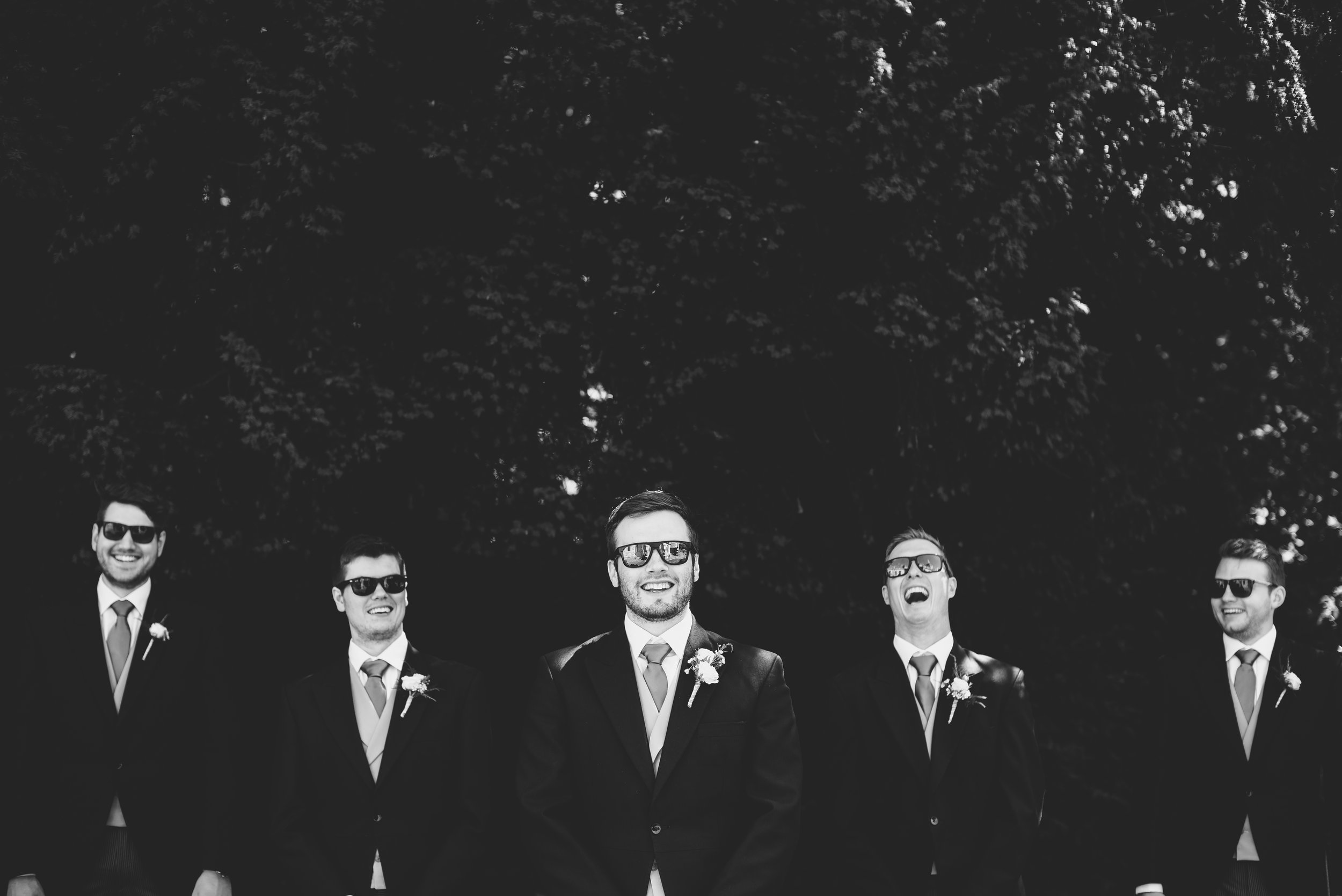 Group of five men in suits, with three wearing sunglasses, standing outdoors in front of trees; one man in the center smiling.