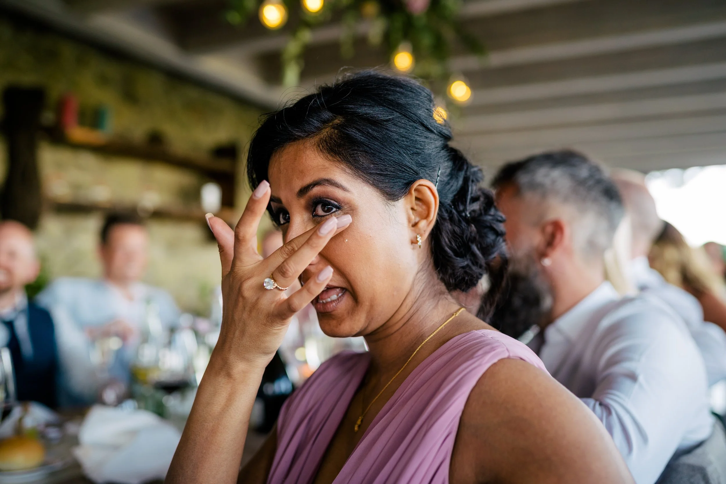 Woman with dark hair and a pink dress wiping tears while seated at a social gathering with blurred people and string lights in the background.