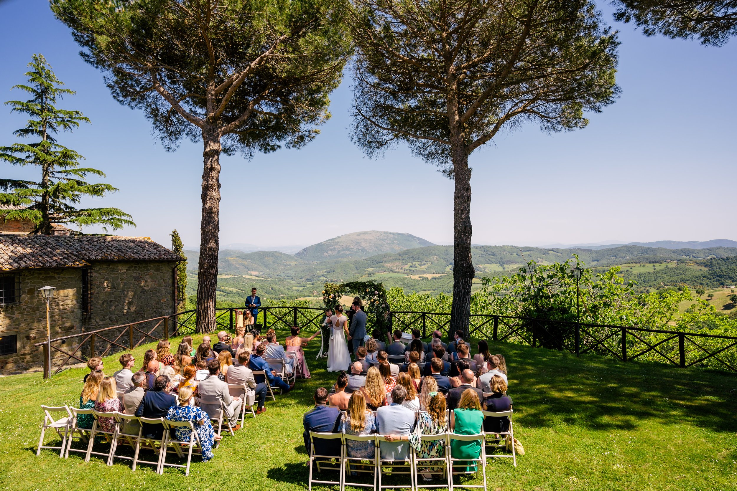 Outdoor wedding ceremony taking place on a grassy lawn with chairs arranged in rows, surrounded by trees and overlooking a scenic landscape of rolling hills and mountains under a clear blue sky.