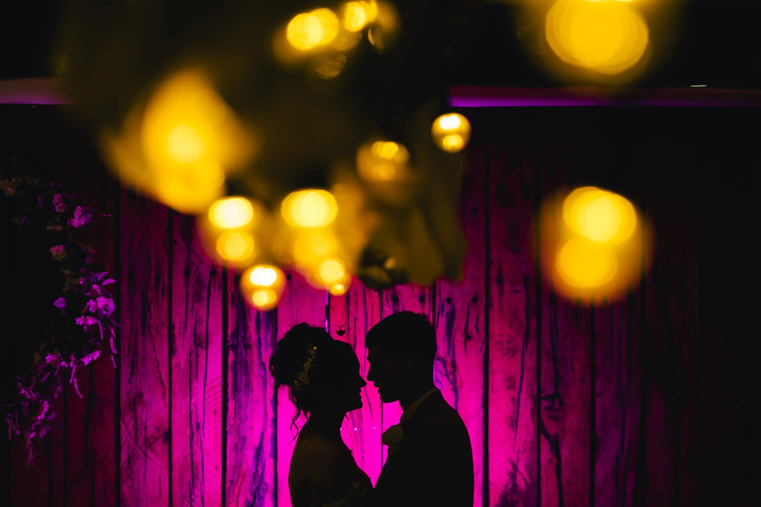 Silhouette of a bride and groom facing each other at their wedding reception with purple lighting background and blurred yellow string lights overhead.