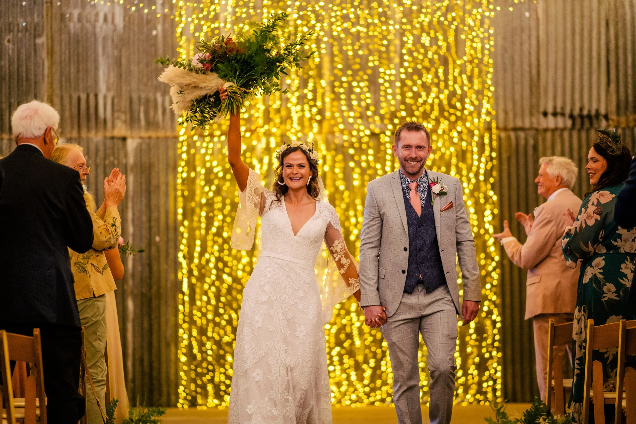 A bride and groom walk hand in hand down the aisle at their wedding ceremony. The bride is holding a bouquet of flowers overhead and smiling. The background is decorated with warm yellow string lights, creating a festive atmosphere.