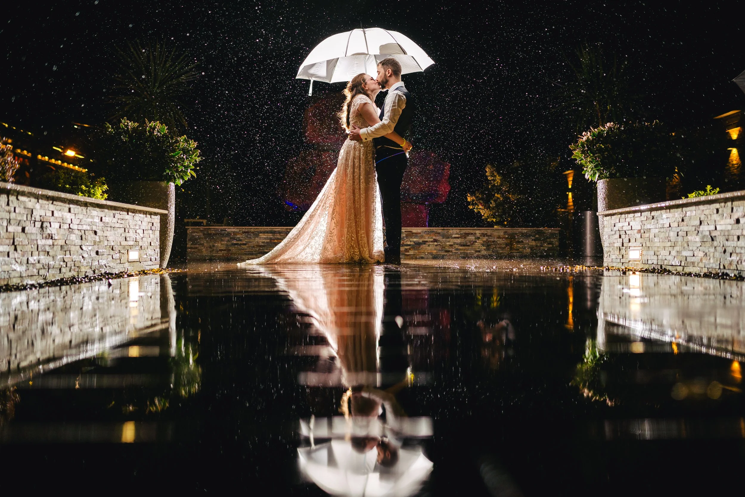 Couple in wedding attire kissing under an umbrella at night, rain falling, with their reflection visible in a puddle, lit by nearby lights.