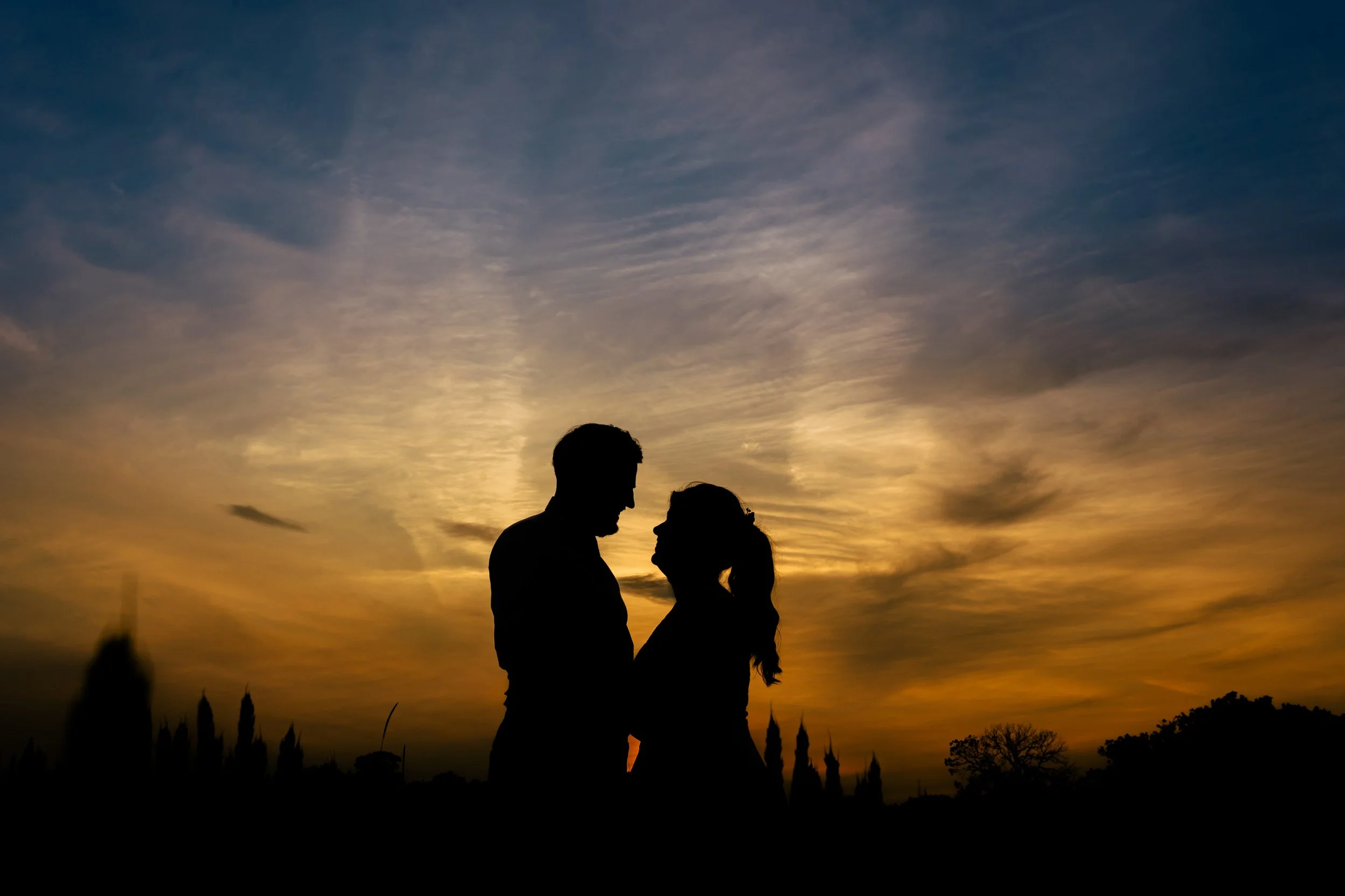 Silhouette of a couple standing close together during a sunset, with the sky painted in warm orange and yellow hues.