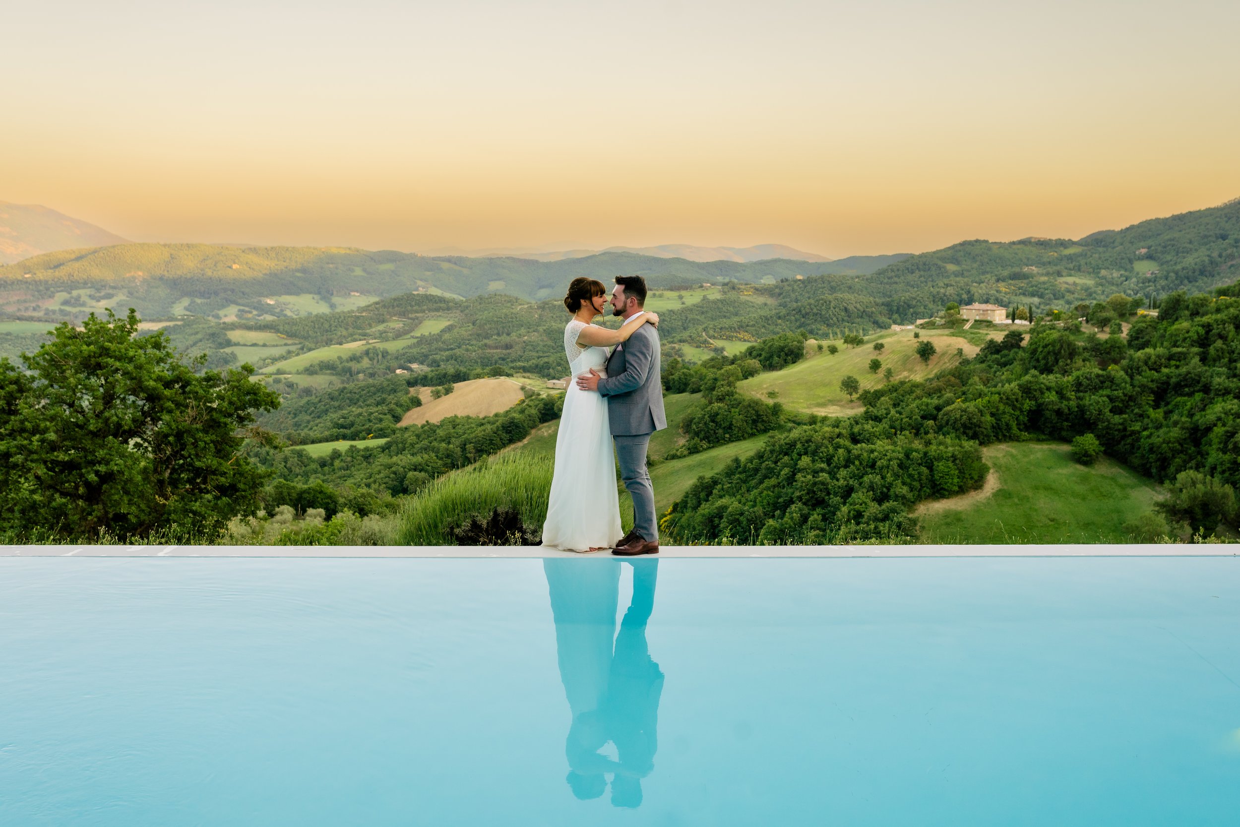 A bride and groom standing in an infinity pool, embracing with a scenic green landscape and hills at sunset in the background.