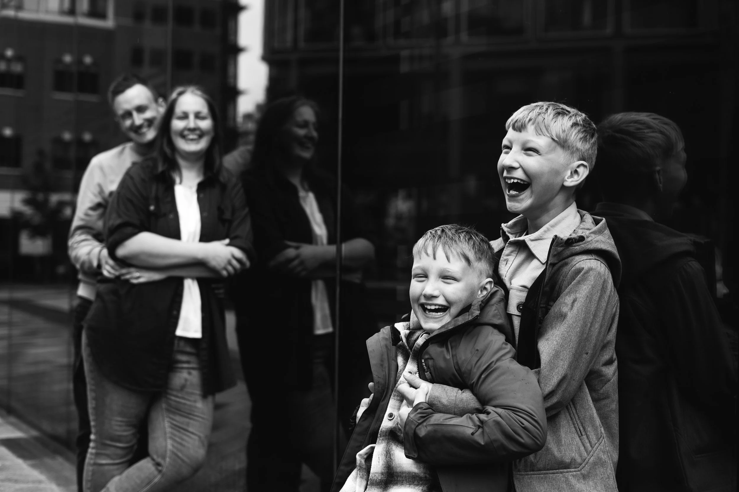 Two young boys laughing and hugging in front of a reflective glass wall, with three adults smiling in the background.