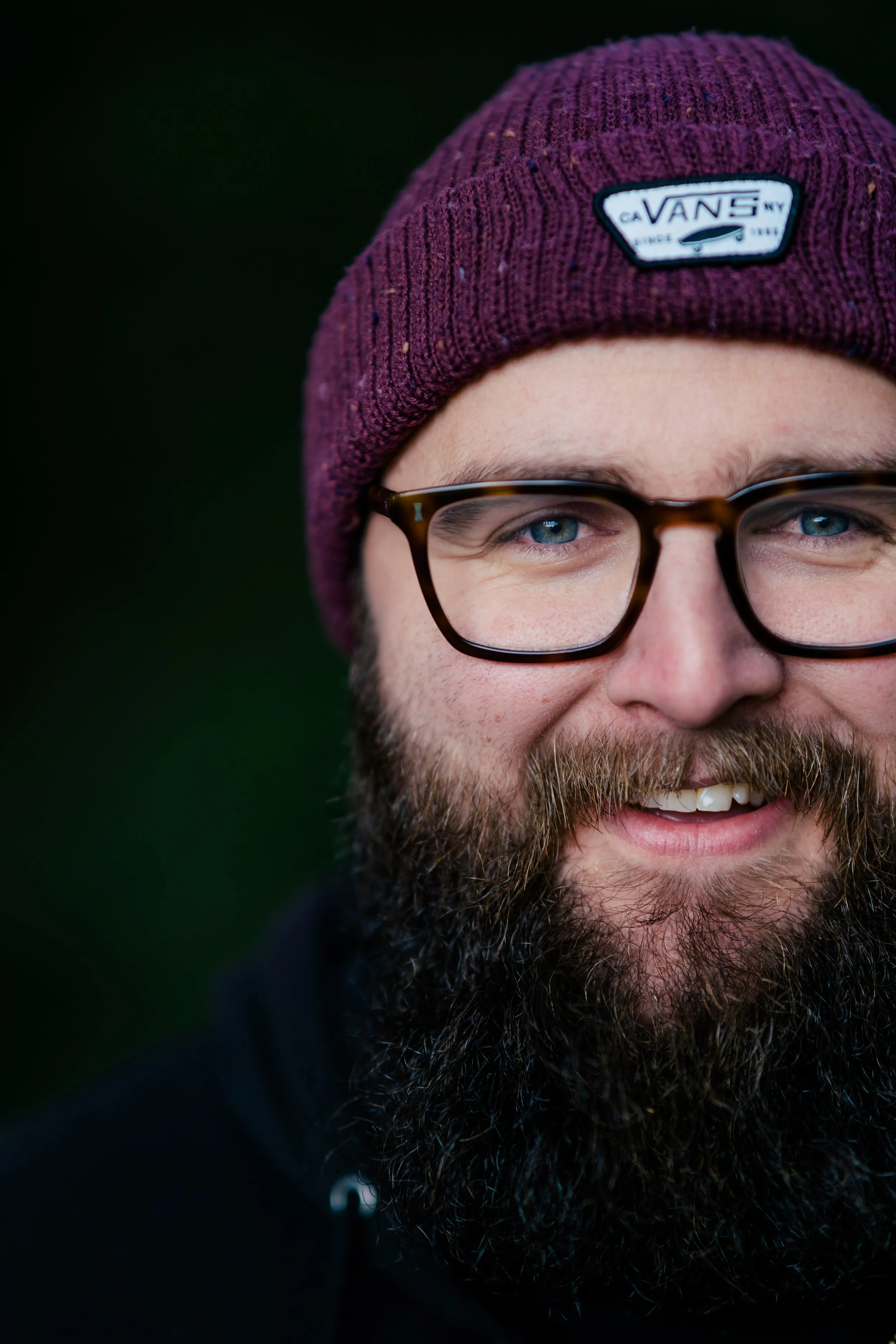 Close-up of a smiling man with a full beard, glasses, wearing a maroon knit beanie with a Vans logo, against a dark blurred background.