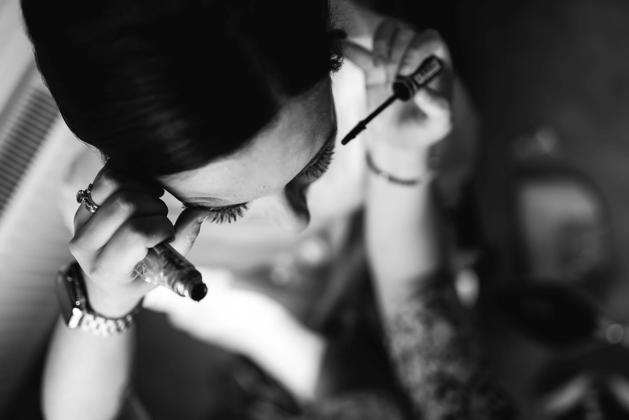 A woman with dark hair applying mascara while looking into a mirror.