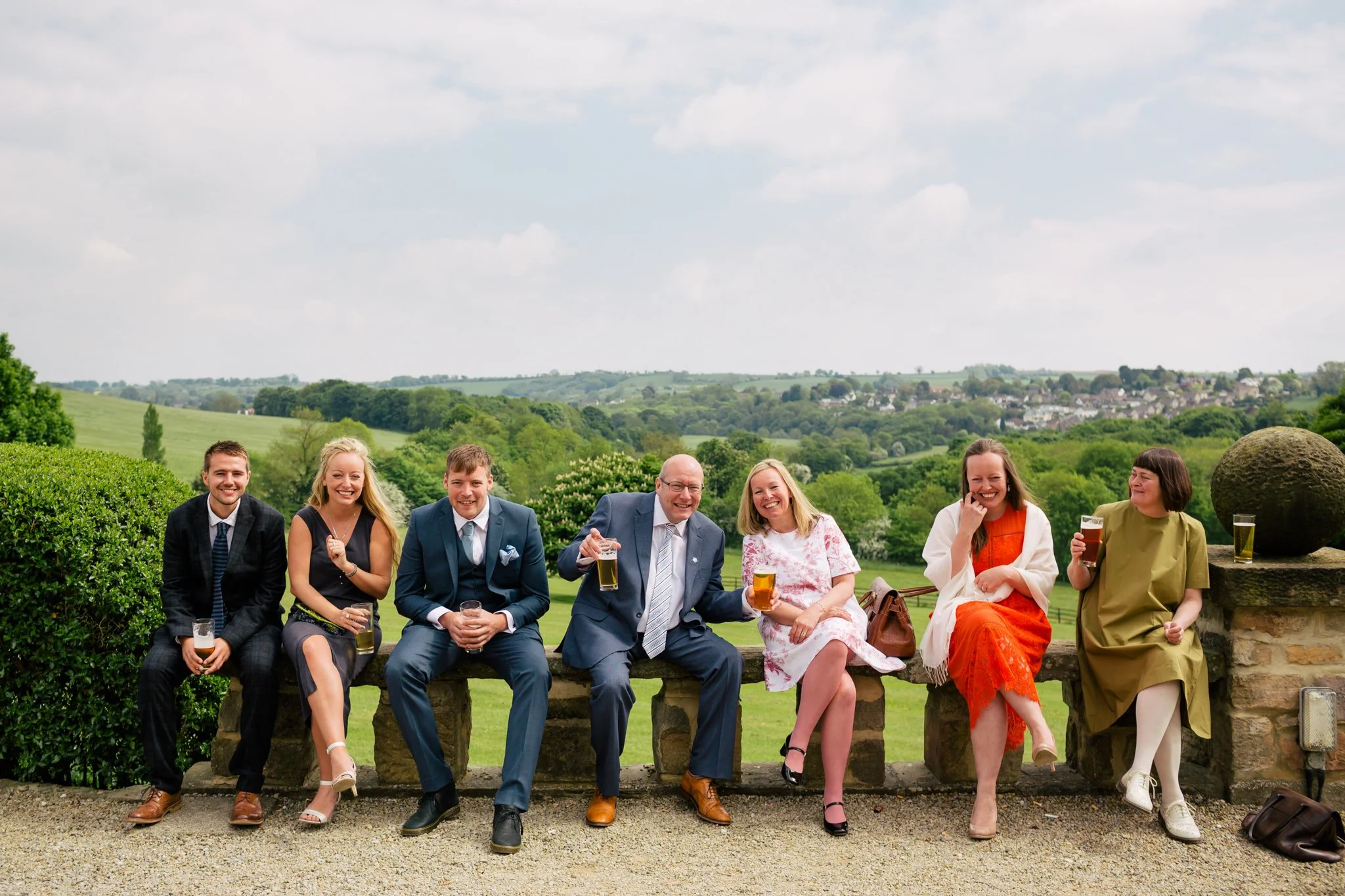 Group of seven people sitting on a stone ledge outdoors, holding drinks and smiling, with green hills and trees in the background.