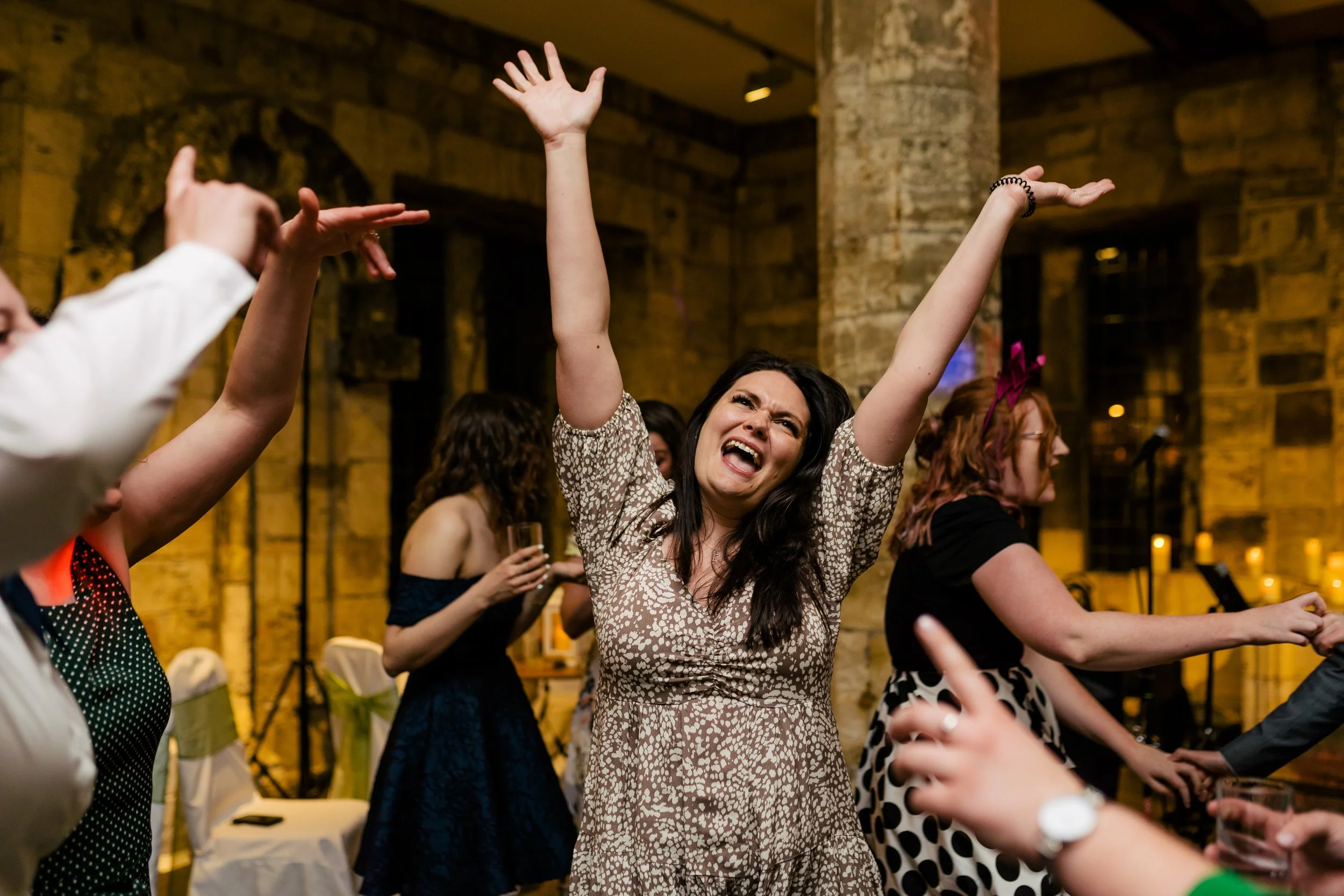 Woman with dark hair in a patterned dress dancing and singing with arms raised at a party or celebration, surrounded by other women and guests in a warmly lit indoor venue.