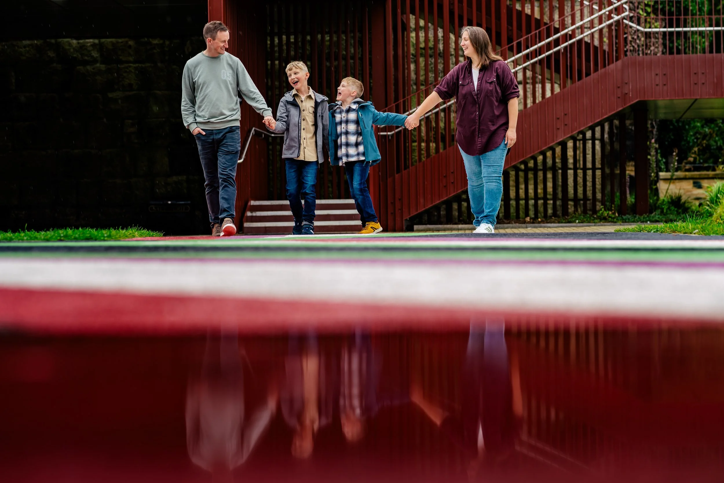 A family of four, two adults and two children, are holding hands and walking together outside on a cloudy day, with a wooden staircase and railing in the background.