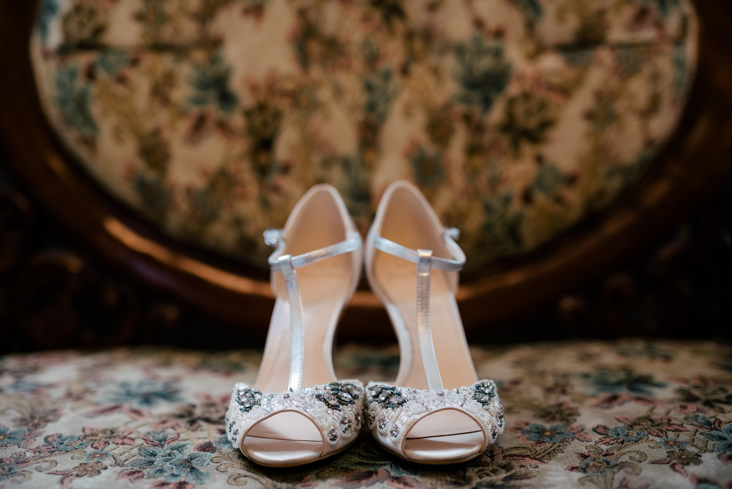 Elegant white wedding shoes with rhinestone embellishments, placed on a floral upholstered surface with a vintage wooden chair in the background.
