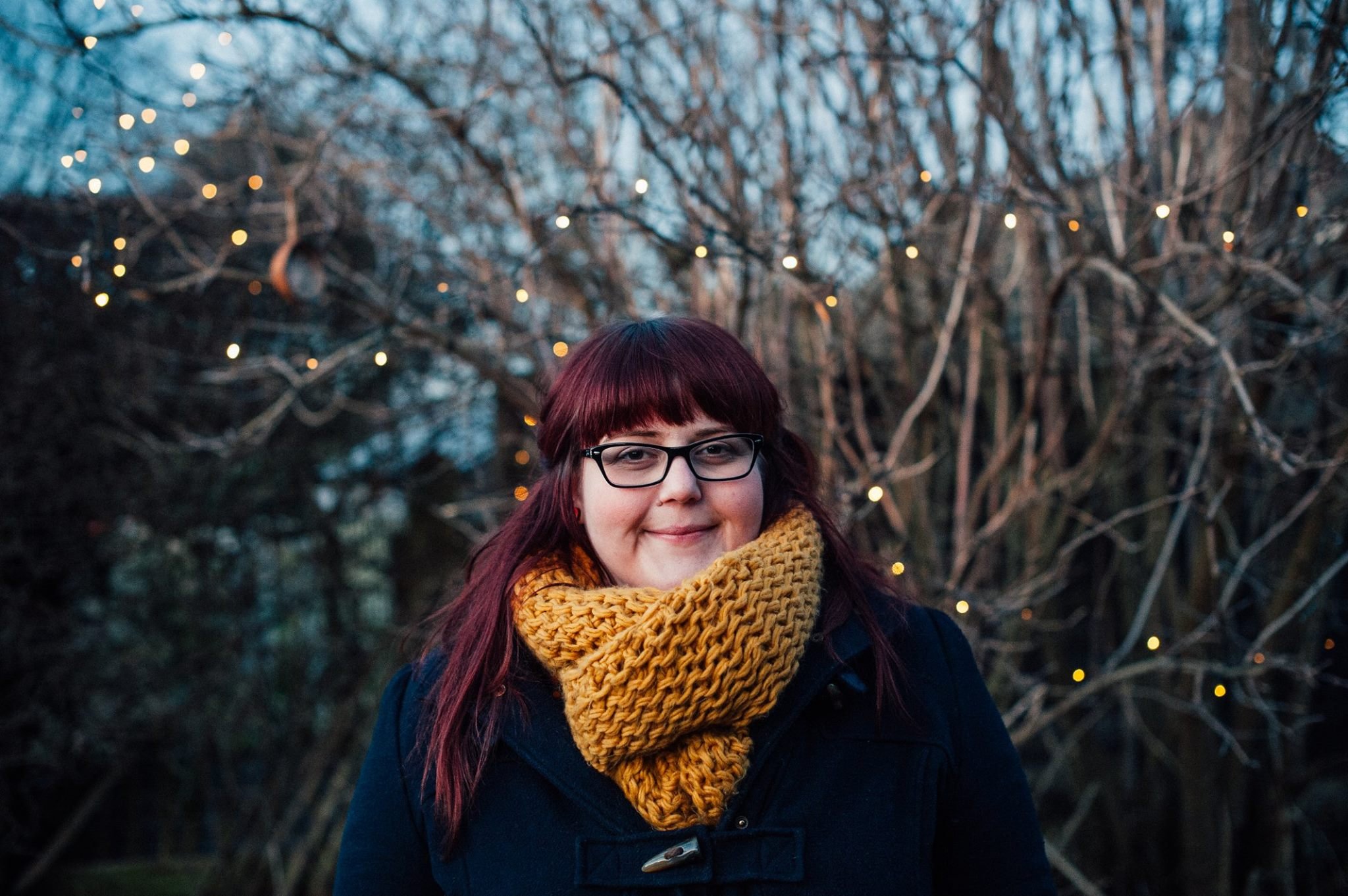 A young woman with red hair, glasses, and a yellow knit scarf smiling outdoors in front of tree branches with small string lights.