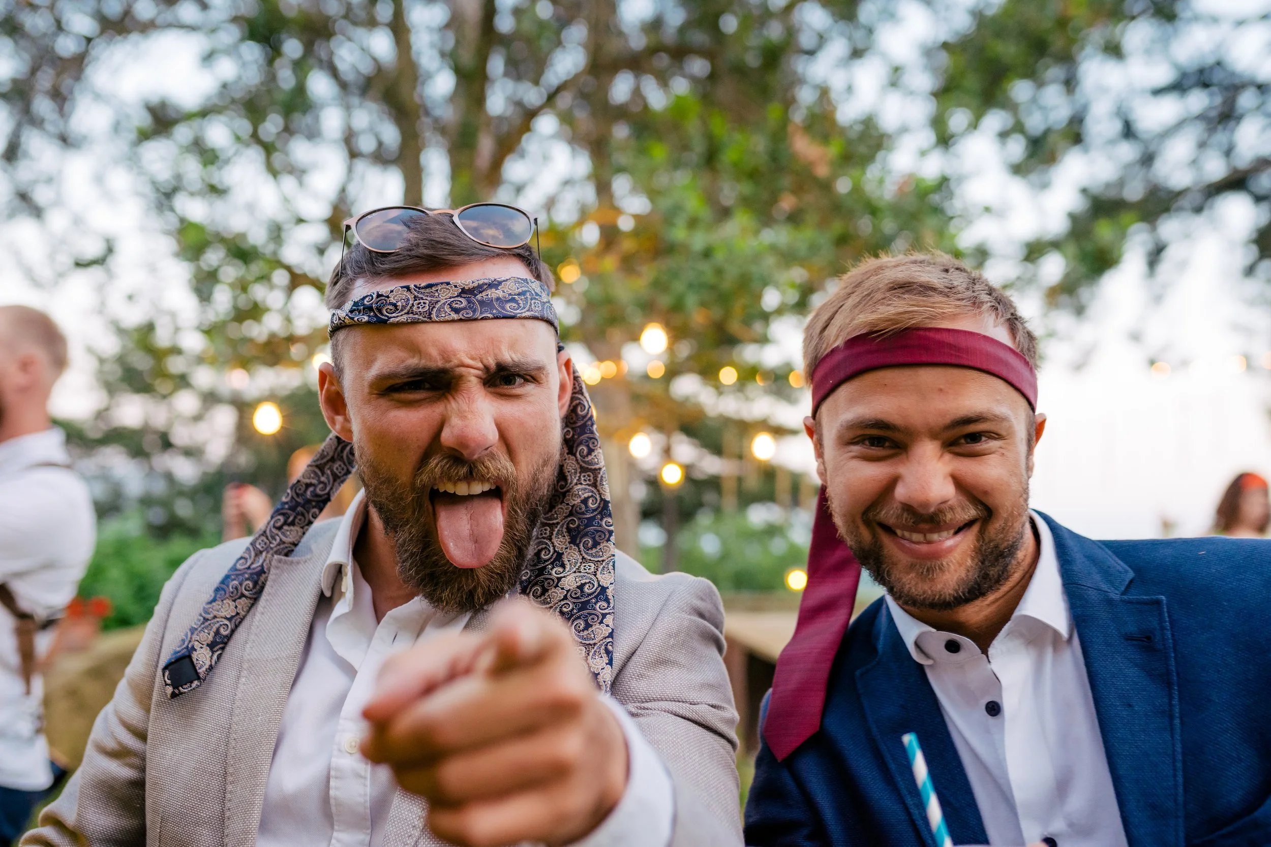 Two men wearing headbands at an outdoor party, one pointing at the camera and sticking out his tongue, while the other smiles. There are trees and blurred lights in the background.