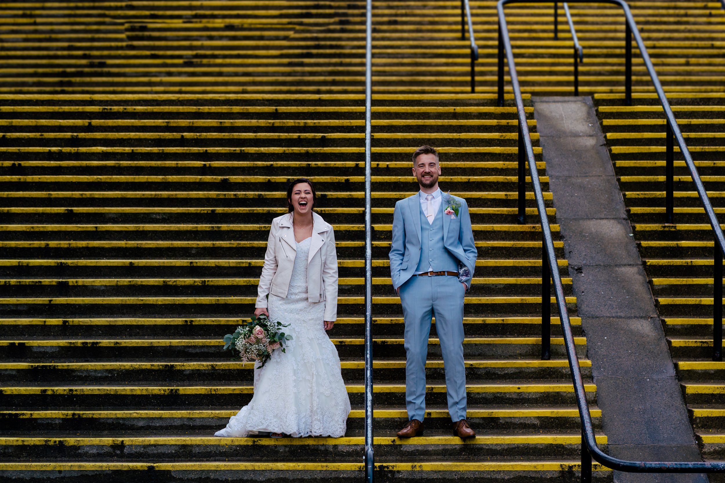 A newlywed couple standing on yellow and black striped stairs, the bride in a white wedding dress holding a bouquet, and the groom in a light blue suit with a boutonniere, both smiling and laughing.