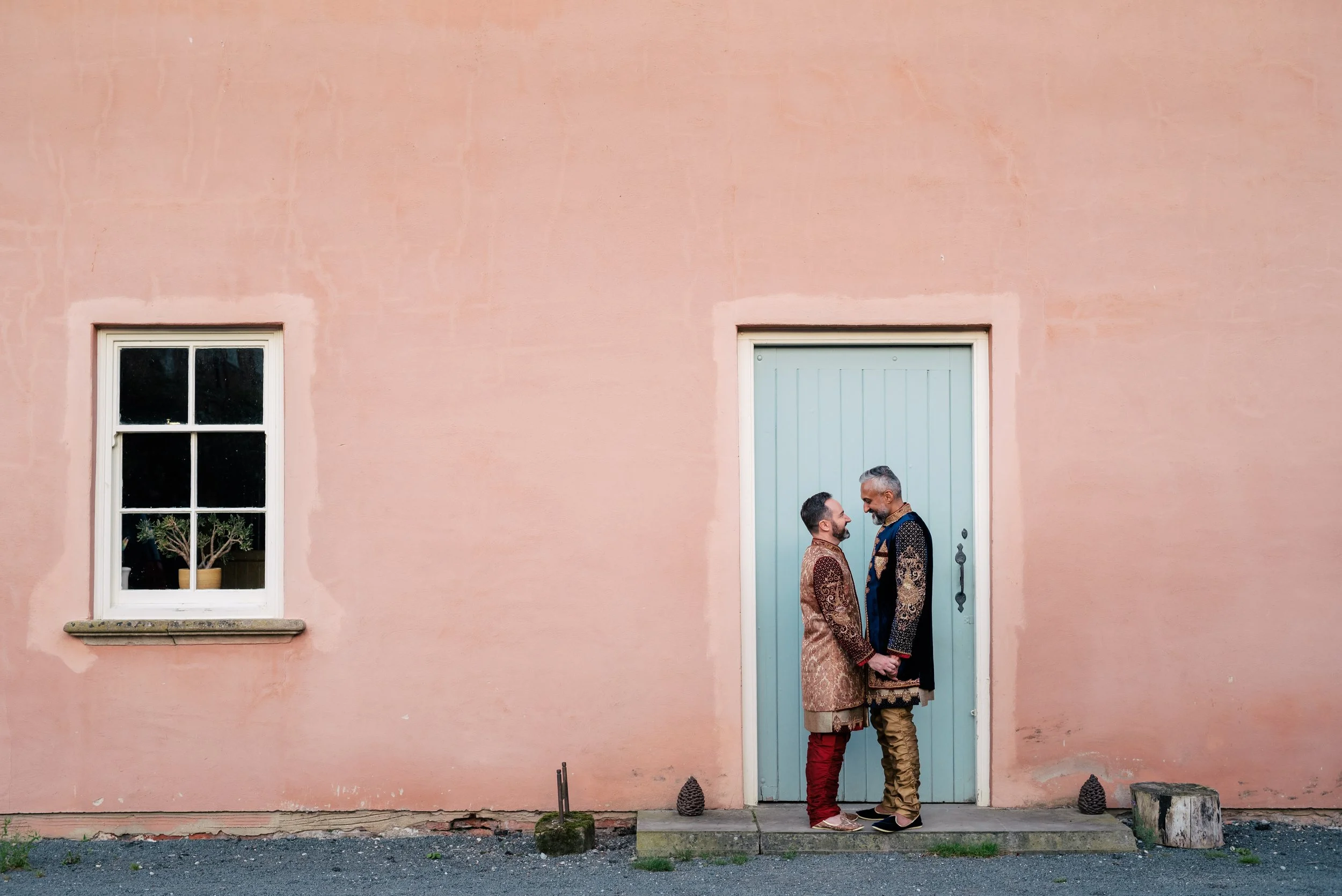 Two men in traditional Indian attire holding hands and smiling at each other in front of a pink wall with a window and a blue door.