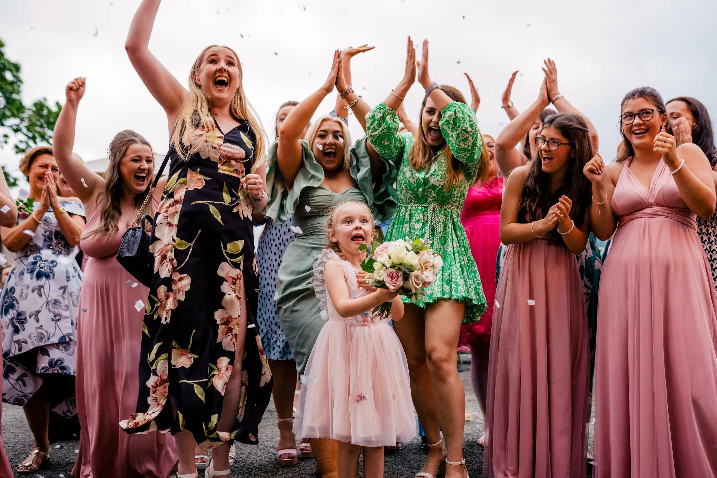 A group of women and girls celebrating outdoors, with one young girl in a pink dress holding a bouquet of flowers, as confetti falls around them.