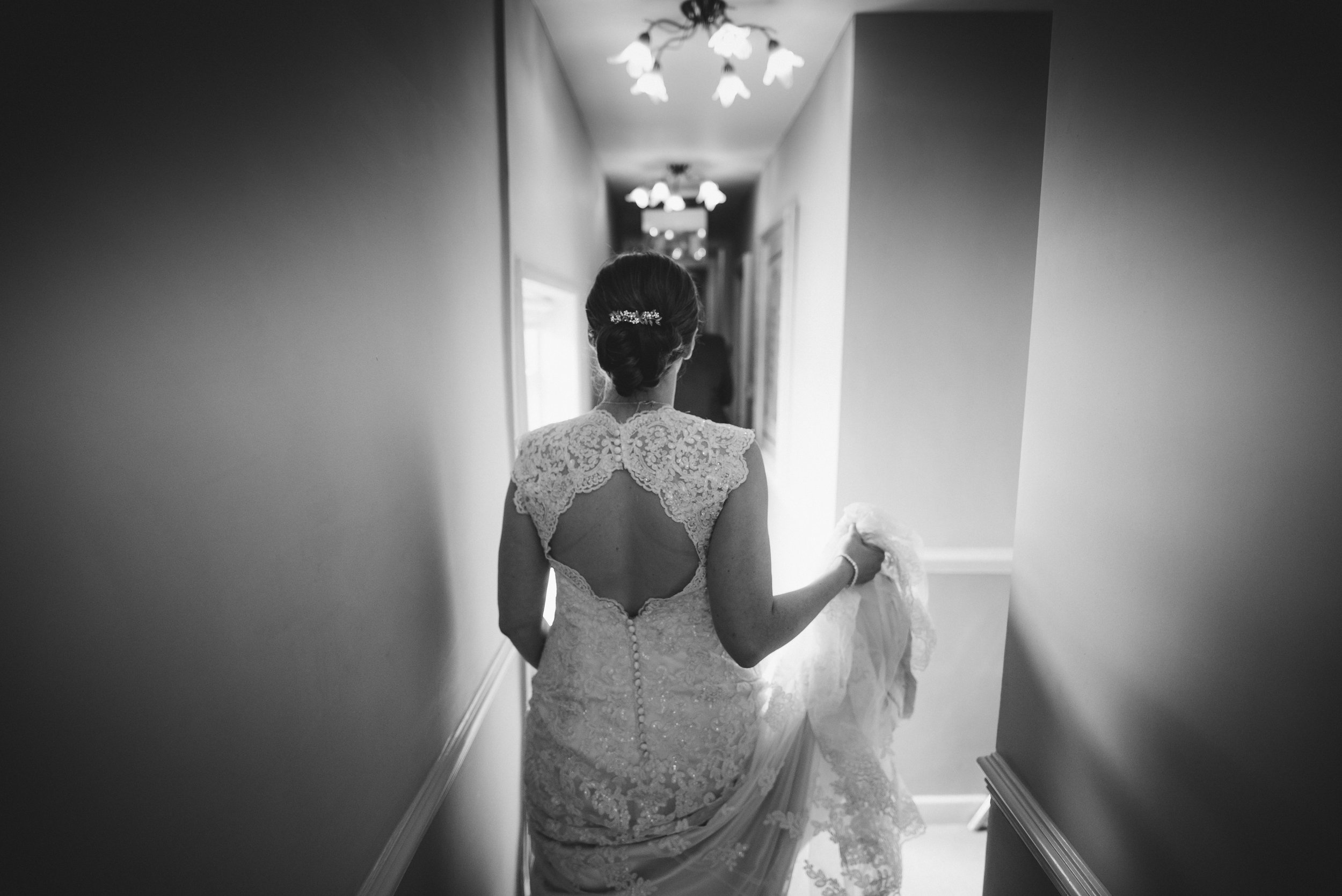 A bride in a wedding dress walking down a narrow hallway, holding her gown, with her back to the camera.