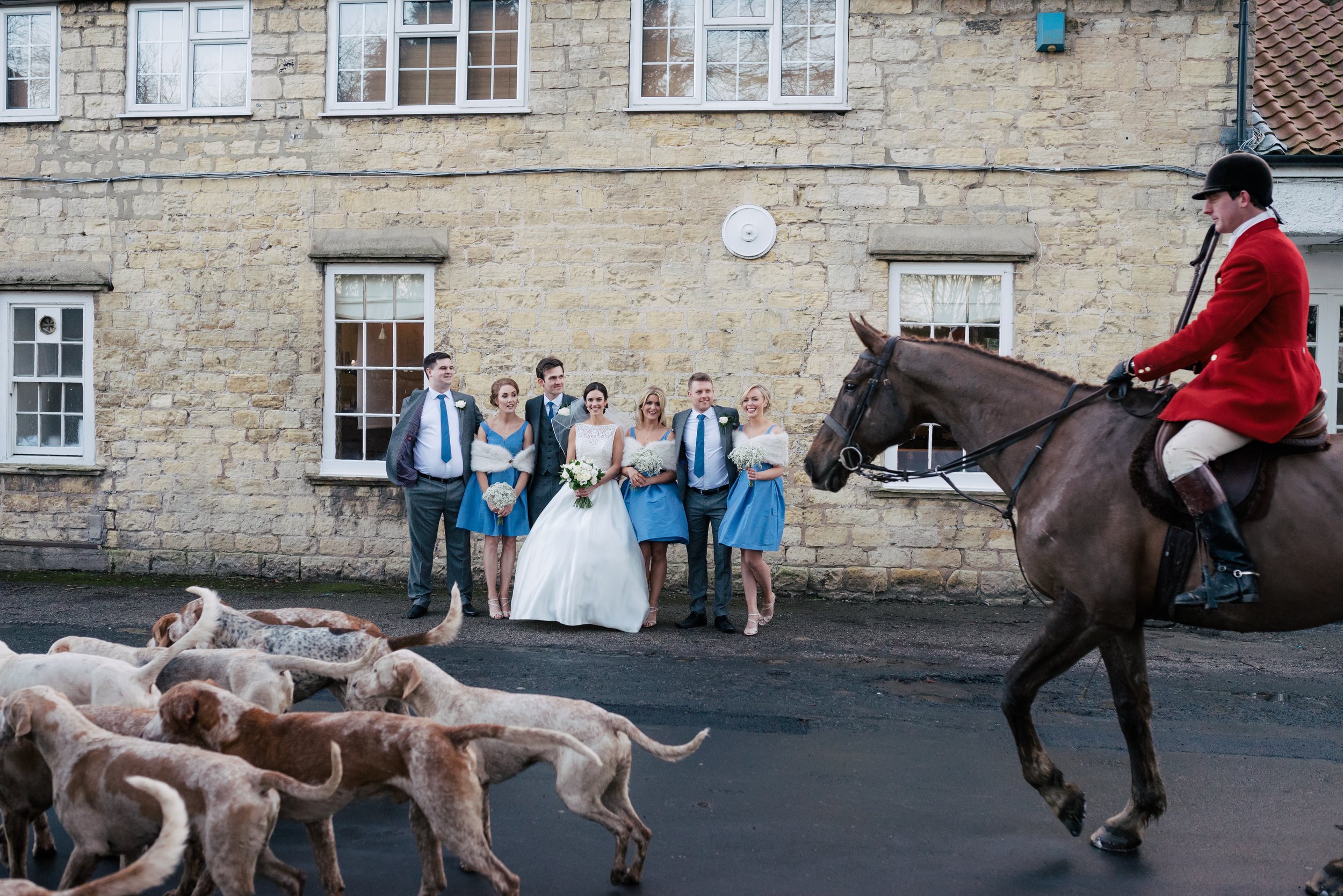 Wedding party standing in front of a stone building, with a groom, bride, and five bridesmaids in blue dresses, and groomsmen, while a person in red riding jacket on a horse approaches with a group of dogs in the foreground.