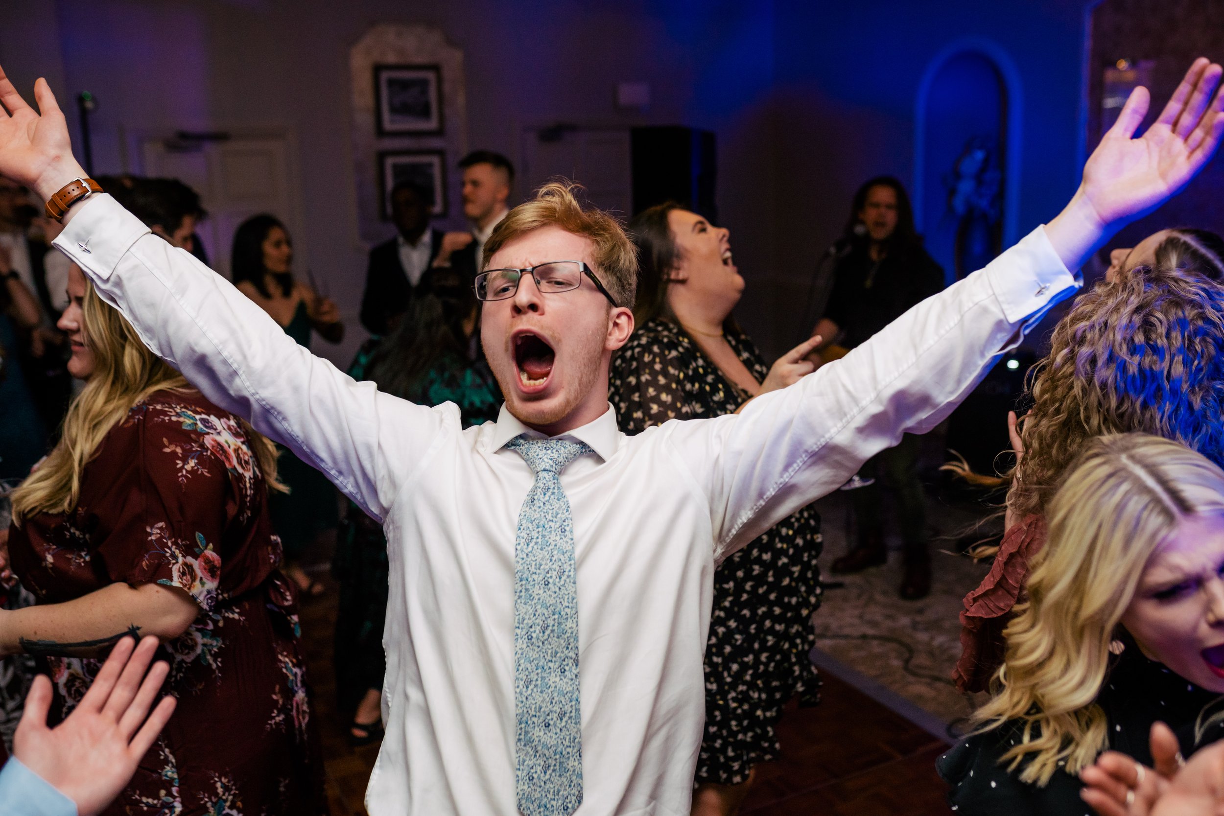 Person with glasses and a white shirt with a patterned tie singing or shouting with arms raised at a lively social event or dance party.