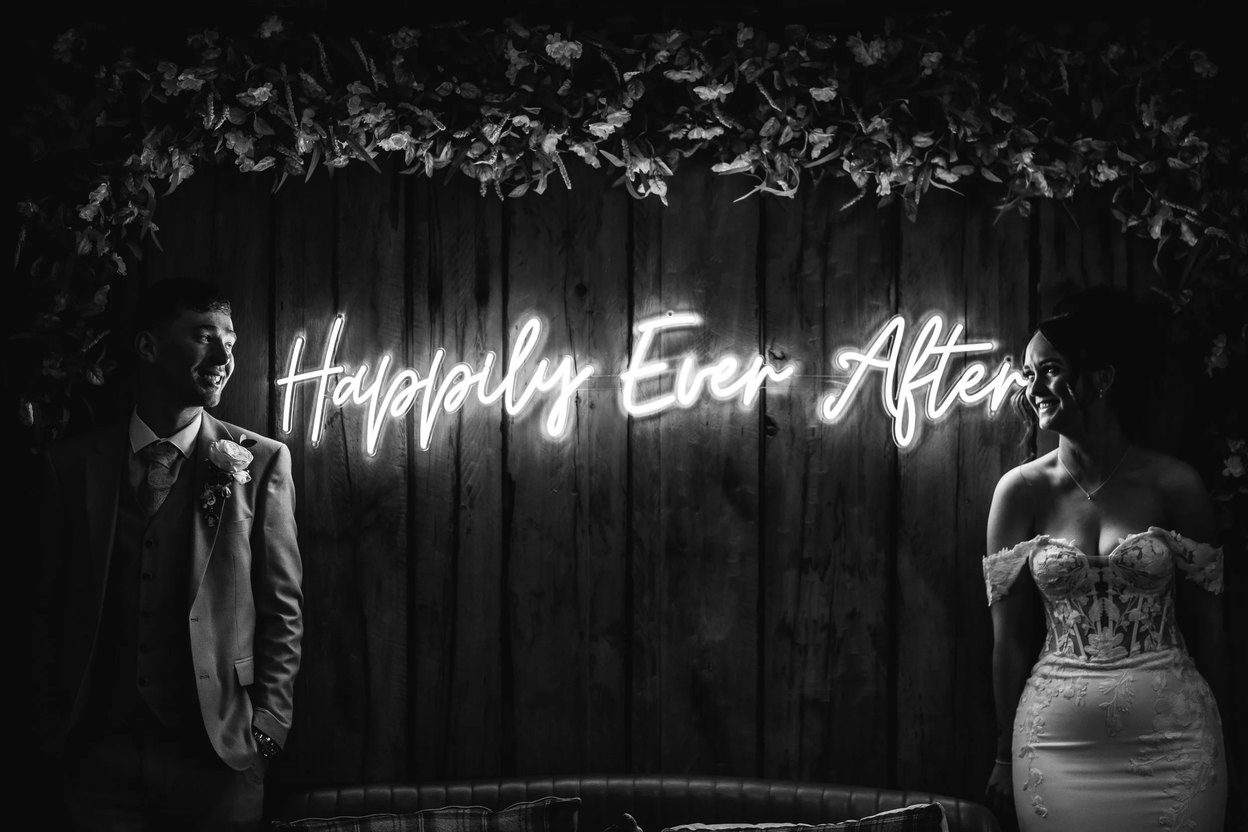A black-and-white photo of a bride and groom standing apart in front of a wooden wall with a neon sign that reads 'Happily Ever After'. The groom is on the left, wearing a suit with a boutonniere, and the bride is on the right, wearing a strapless we