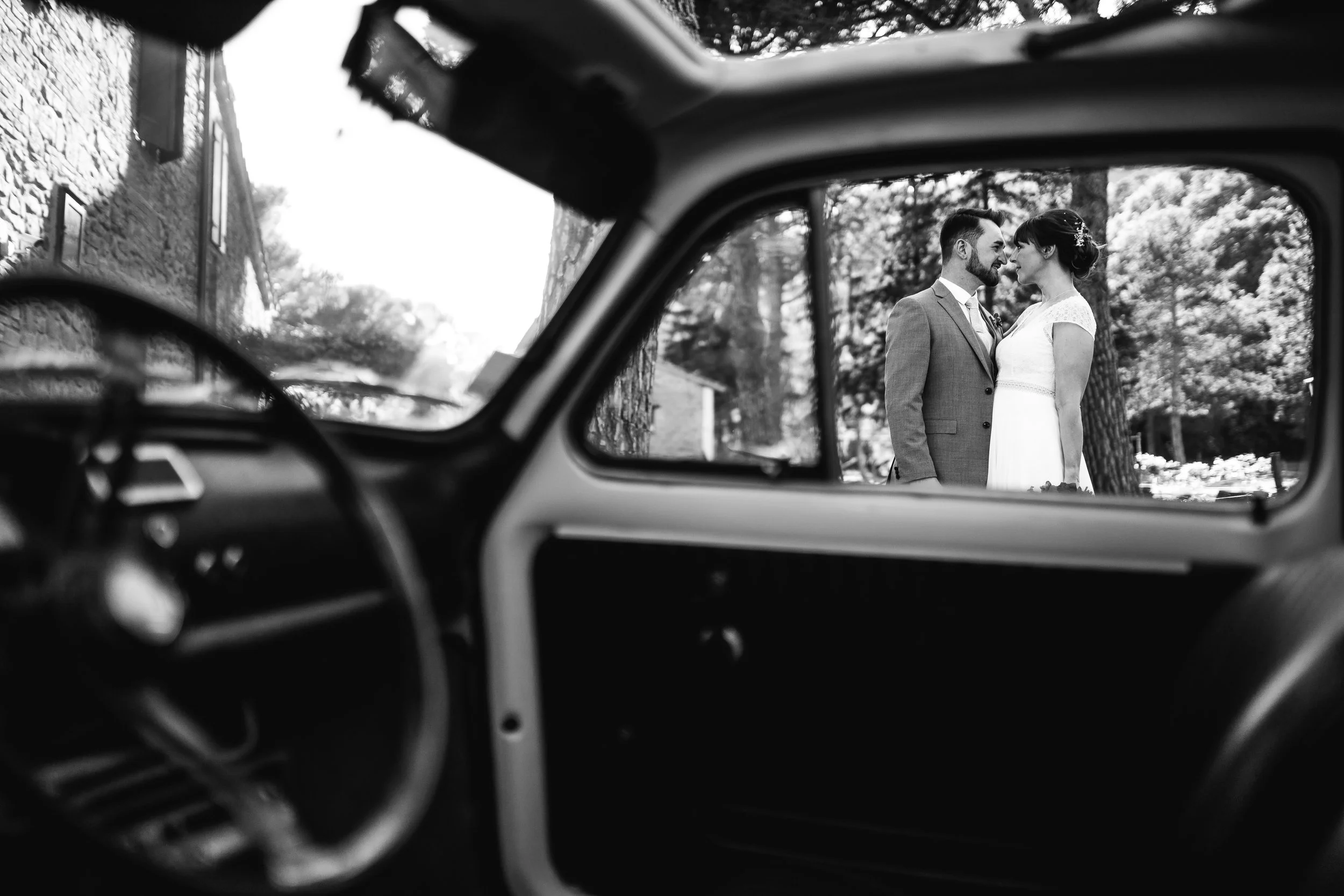 A black and white photo of a happy couple in wedding attire, gazing into each other's eyes, seen through the window of a vintage car.