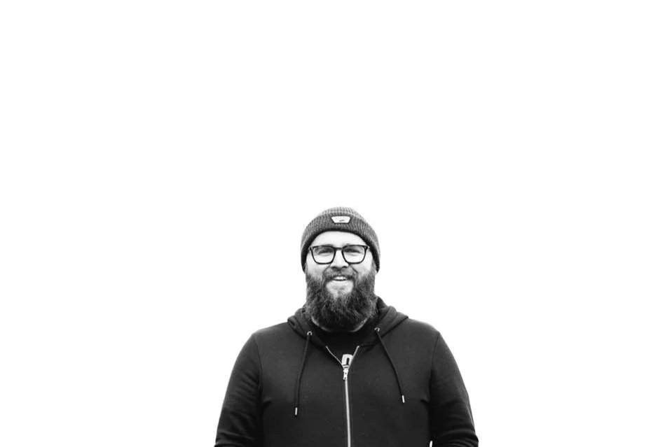 A man with a beard, glasses, and a beanie smiling outdoors, wearing a hoodie.
