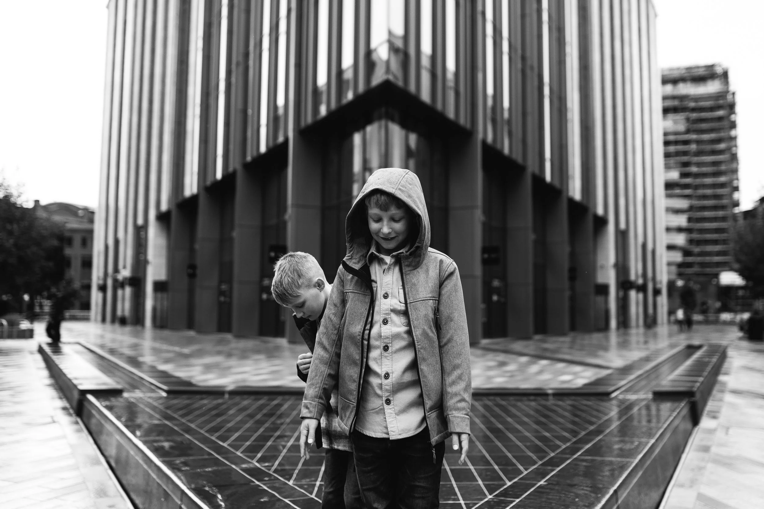 Two children, a girl with a hooded jacket and a boy, standing on a city street in front of a modern building with metallic facade, in black and white.
