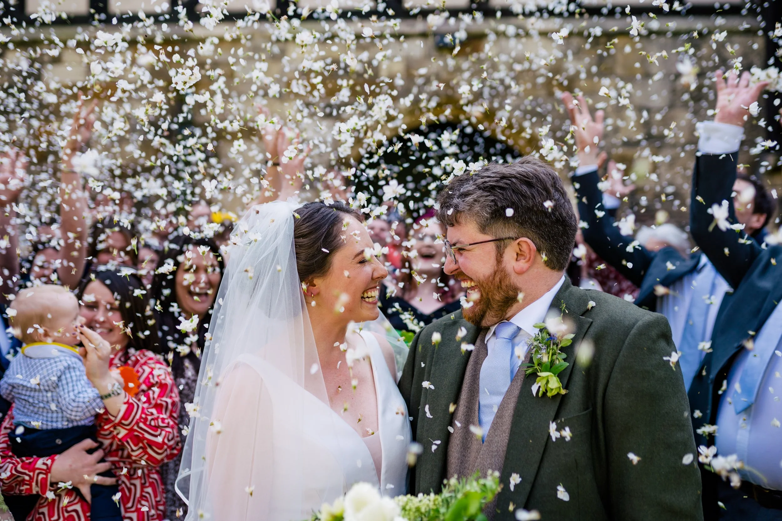A bride and groom smiling at each other surrounded by guests showering them with flower petals outdoors.