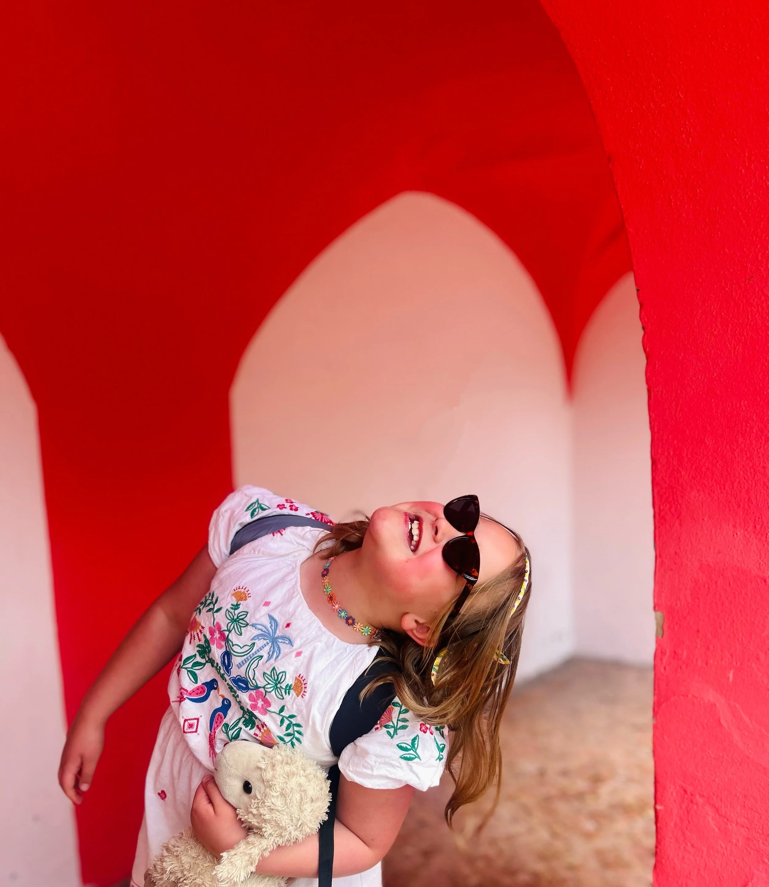 A young girl wearing sunglasses, a white embroidered dress, and a colorful necklace, holding a stuffed animal, smiling and laughing inside an art installation with red and white arched walls.
