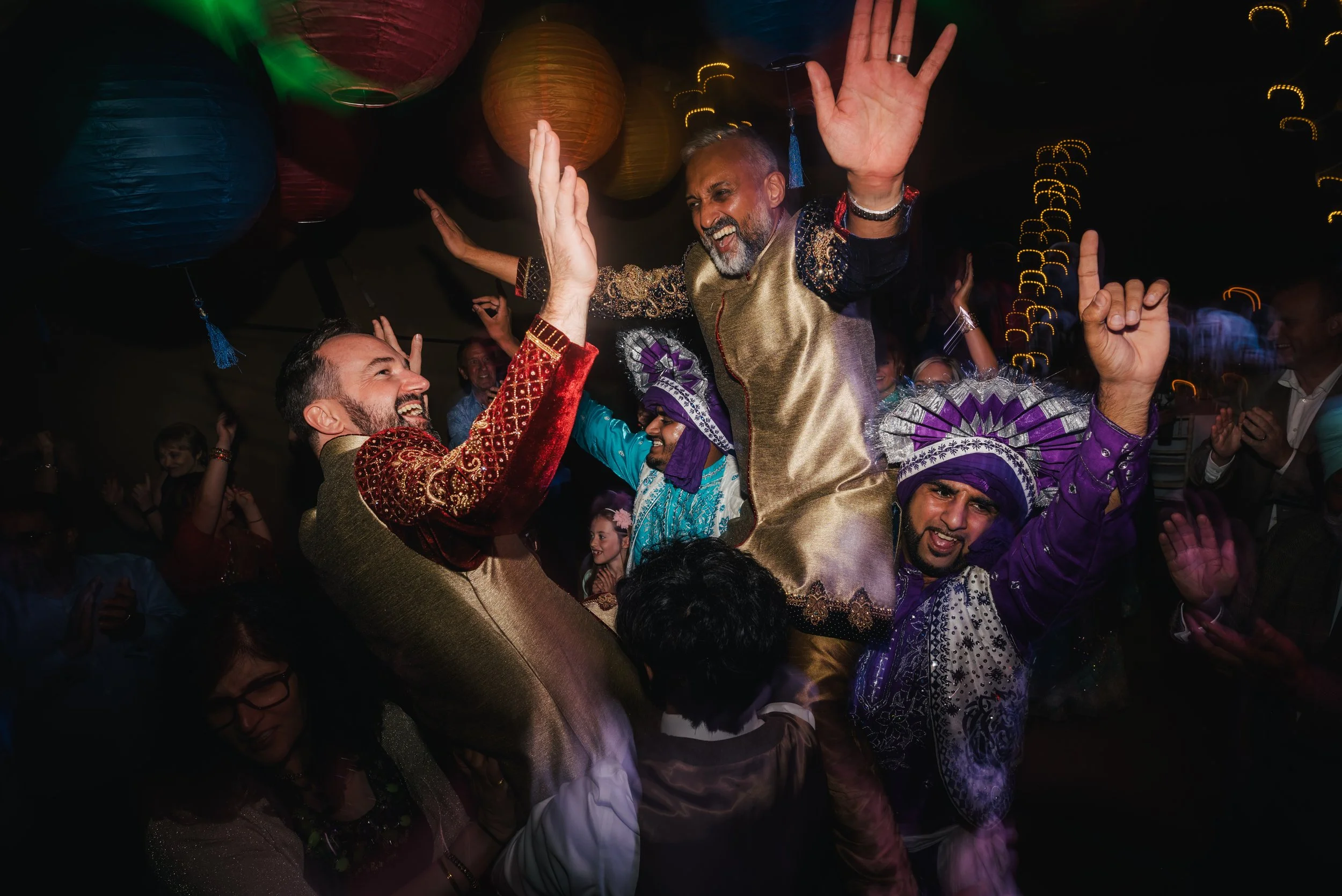 People celebrating at a party, some dressed in traditional South Asian attire, dancing and smiling in a festive atmosphere with colorful lanterns hanging overhead.