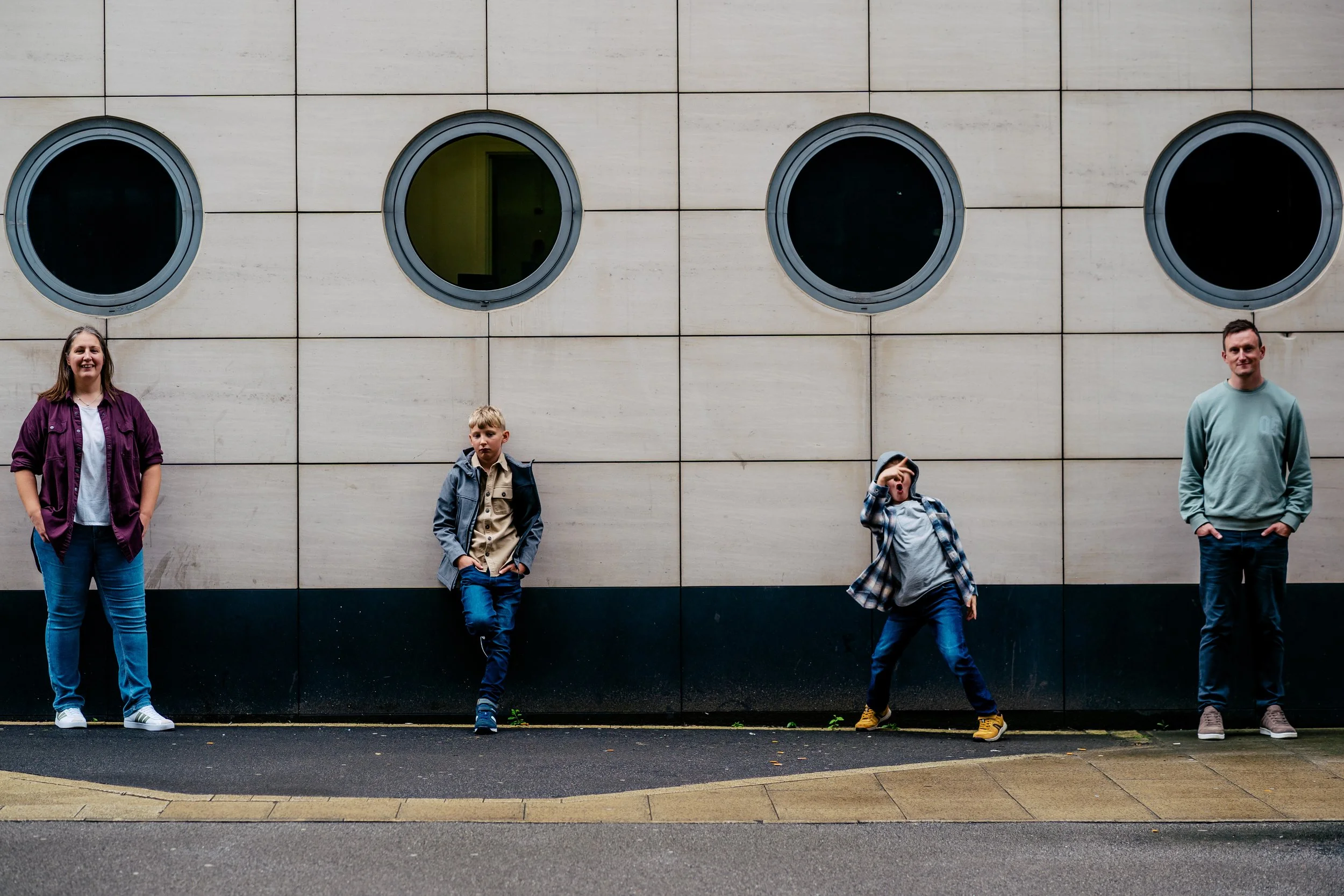 Four people standing against a beige tiled wall with circular windows. From left to right: a woman in a purple jacket and blue jeans, a young boy in a beige shirt and gray jacket, a boy in a plaid shirt and gray hoodie making a gesture, and a man in 