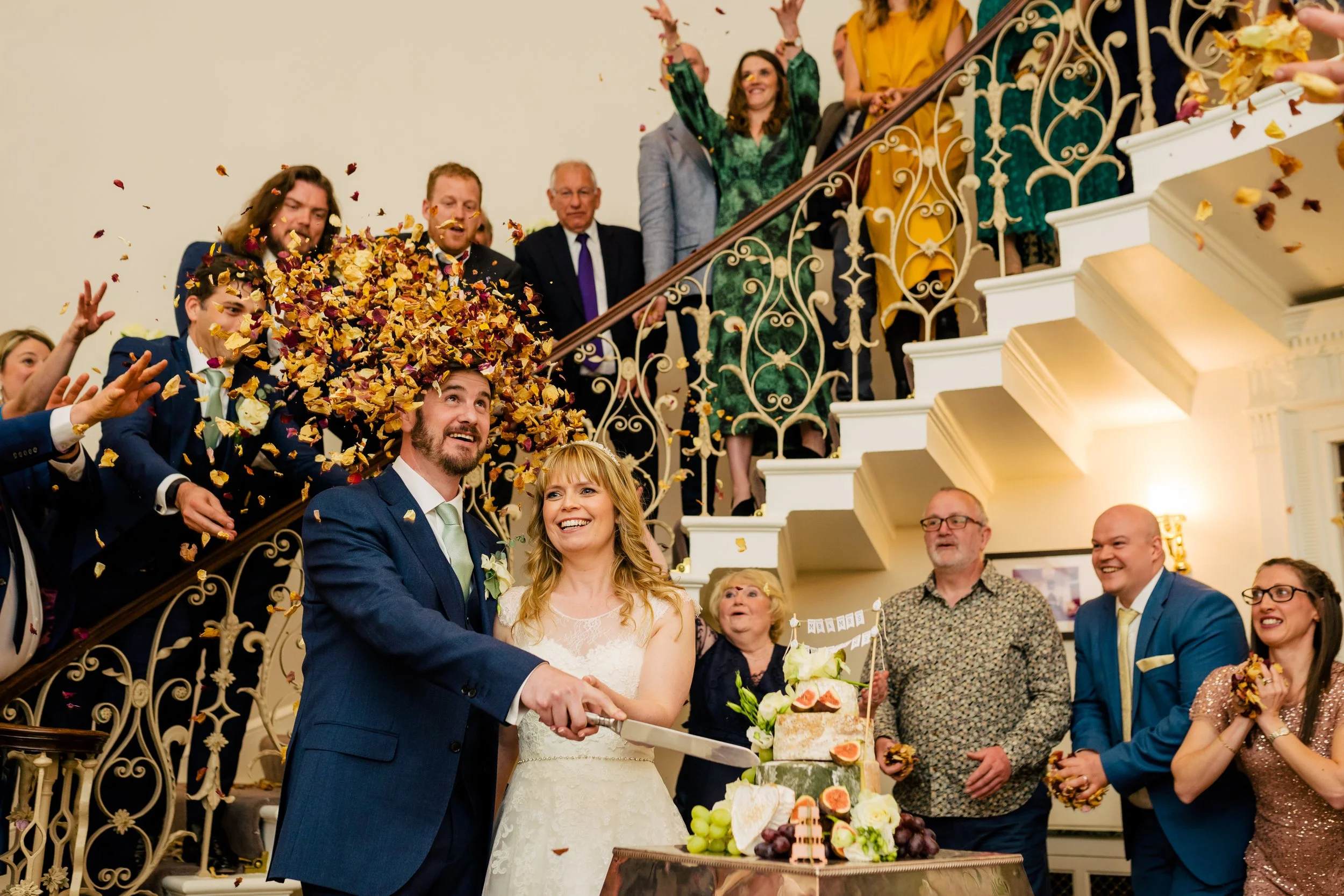 Happy newlywed couple cutting wedding cake while guests throw flower petals overhead, celebrating indoors with ornate staircase and decorations.