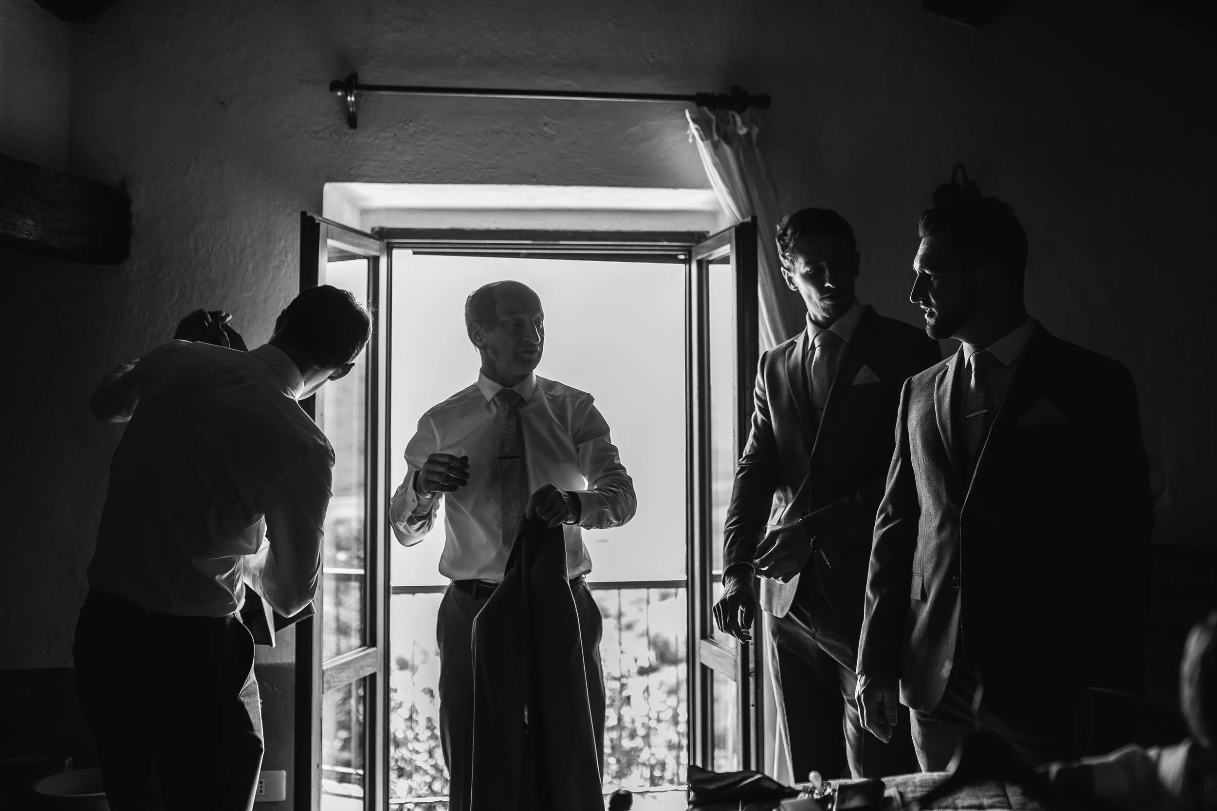 A group of men in formal suits gathered in a room by an open window, with sunlight streaming in and creating silhouette effects.