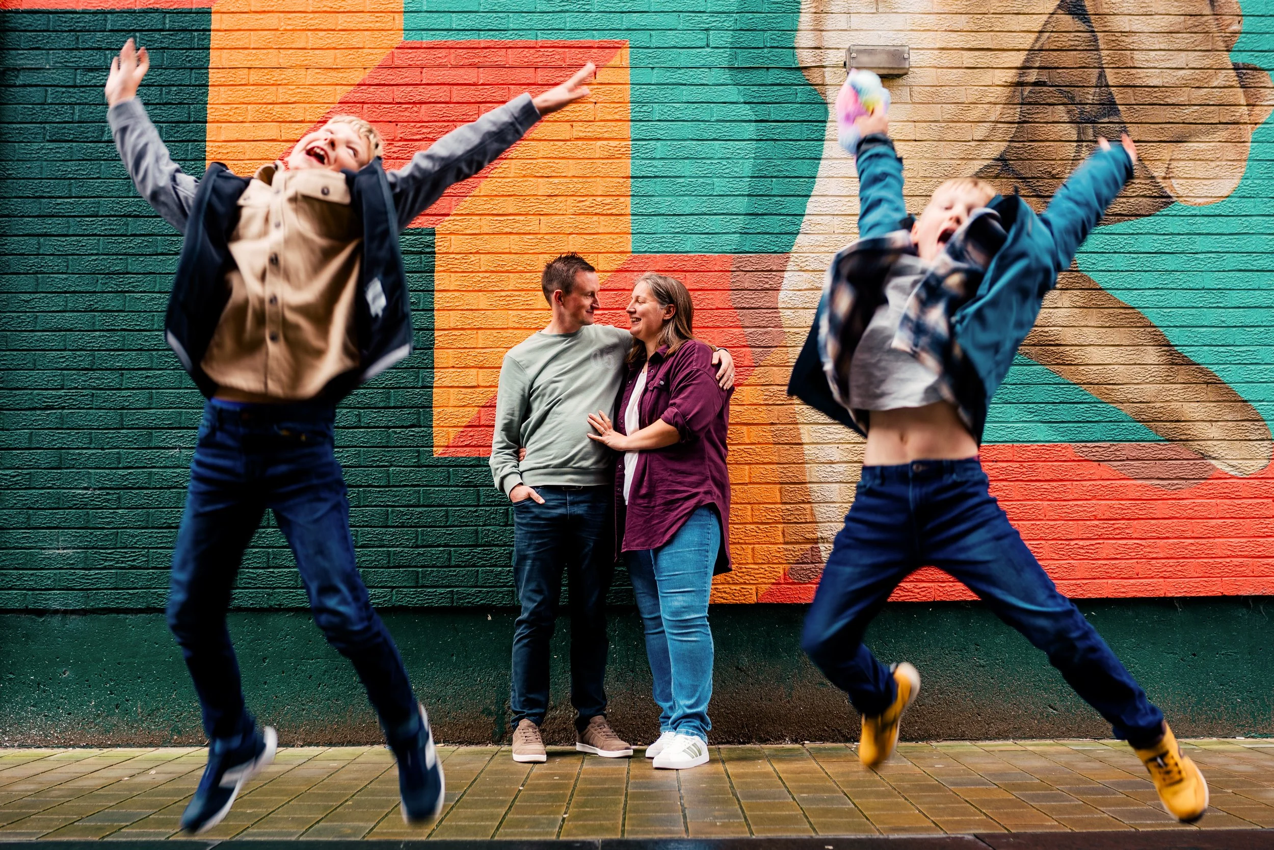 Two children jumping in the air in front of a colorful mural, with a smiling couple standing close together and looking at each other in the background.