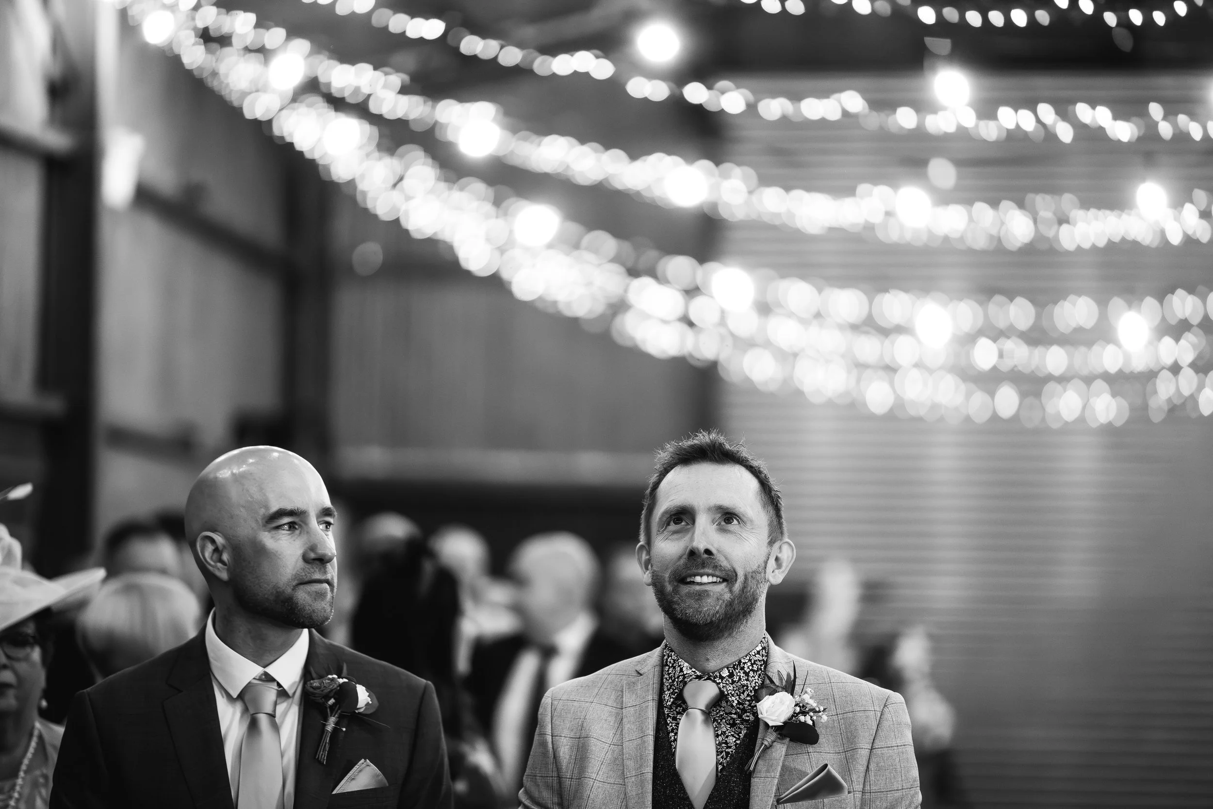 A wedding ceremony with two grooms, one with a shaved head and beard, and the other with short hair and a beard, standing indoors decorated with string lights. The groom with short hair is smiling, and both are dressed in suits with boutonnières.