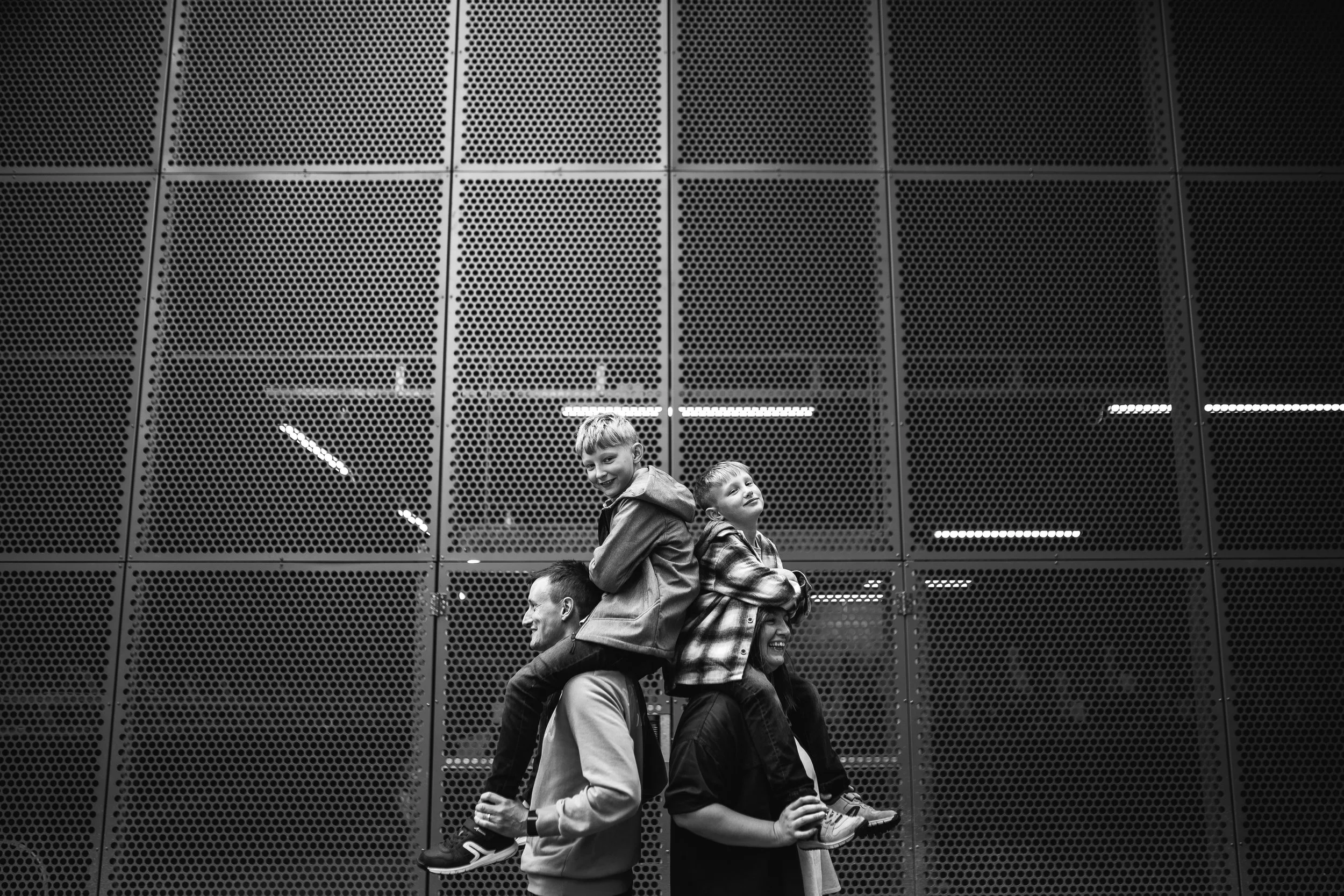 Two adults carrying two children on their shoulders in front of a perforated metal wall, black and white photo