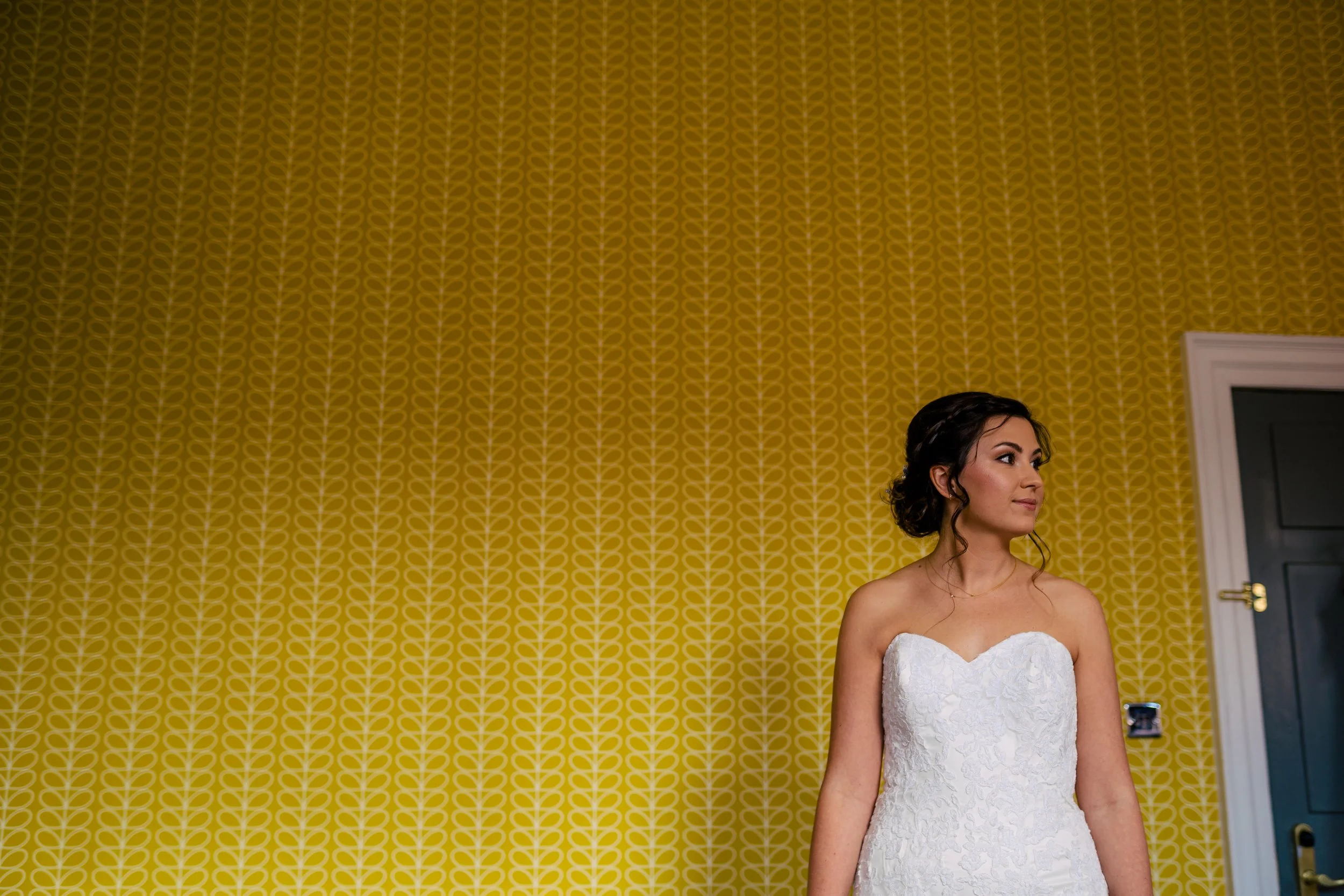 A woman in a strapless white wedding dress standing in front of a yellow patterned wallpaper.