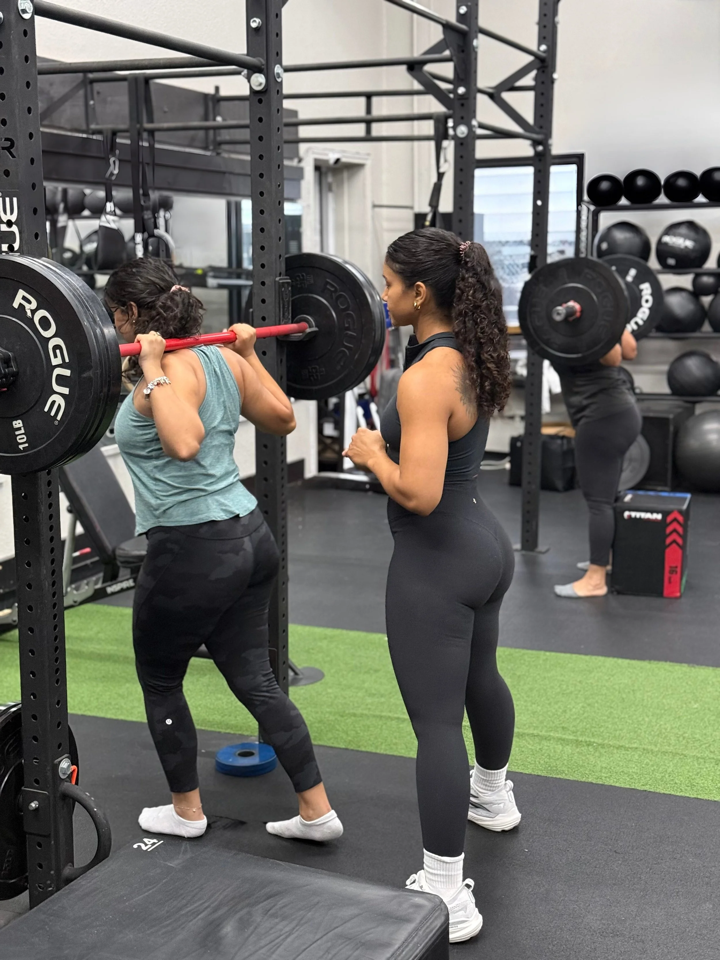 A woman spotting a girl doing a barbell squat in a gym. The girl is holding a barbell with weights on her shoulders, while the woman is observing her. There are shelves with medicine balls and other gym equipment in the background.