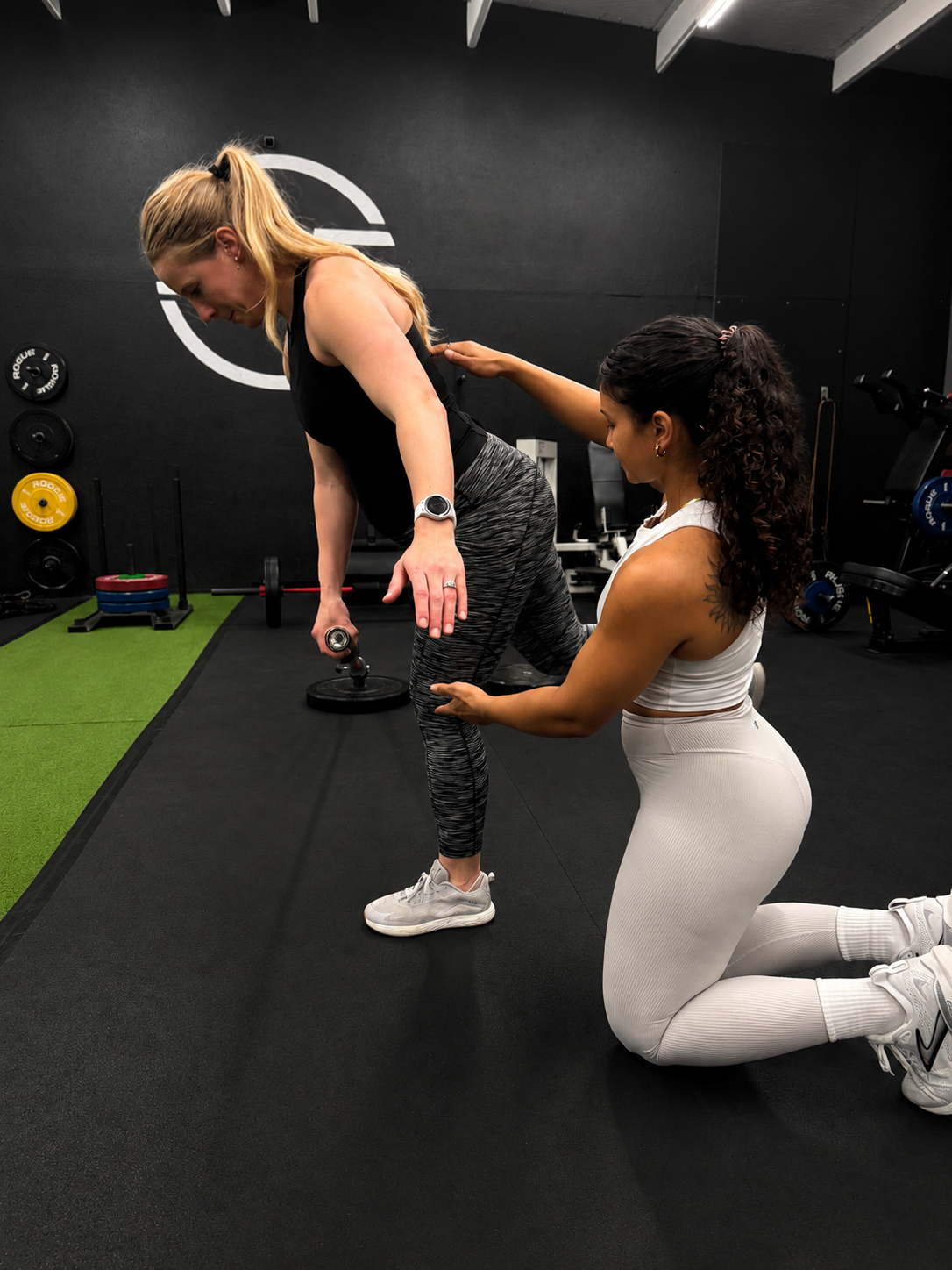 A fitness trainer in white leggings is assisting a pregnant woman in a black top and patterned leggings with a bent-over exercise using dumbbells at a gym.