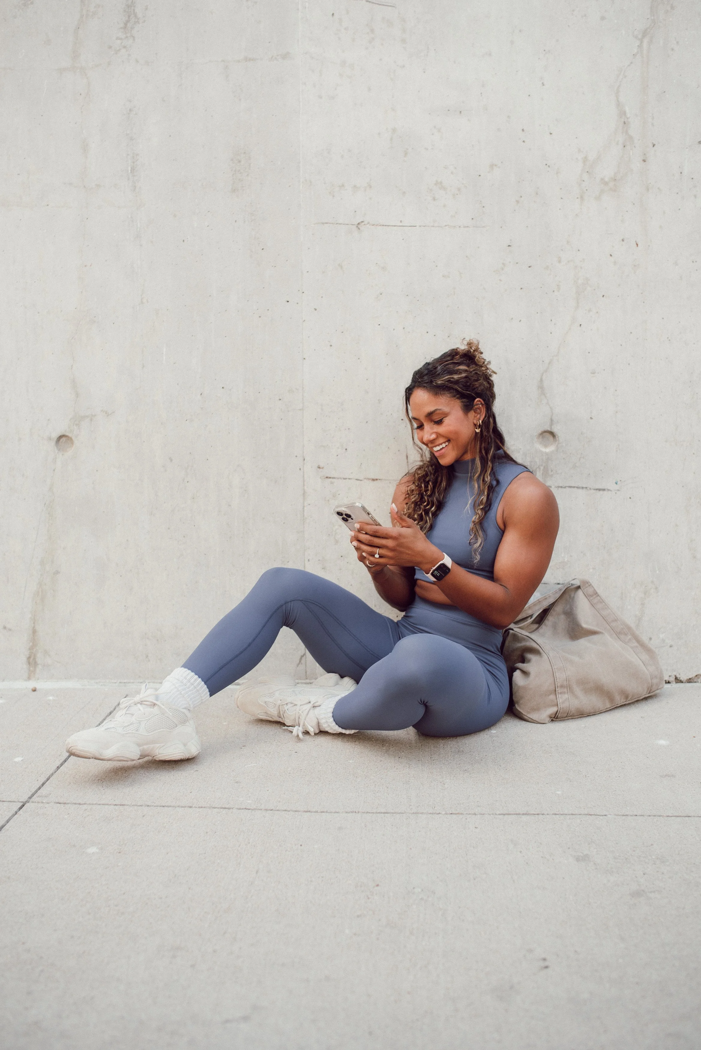 Young woman sitting on the ground, smiling while looking at her phone, wearing workout clothes, in front of a plain concrete wall.