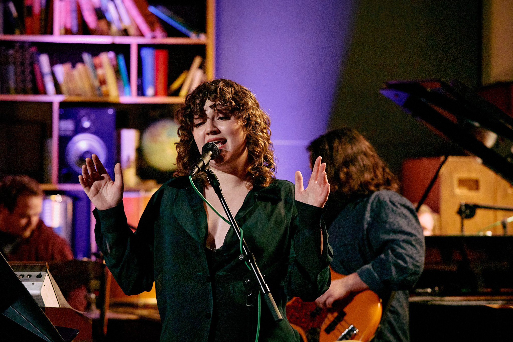 A woman singing into a microphone with eyes closed, in a music studio or performance space, with musicians playing background instruments.