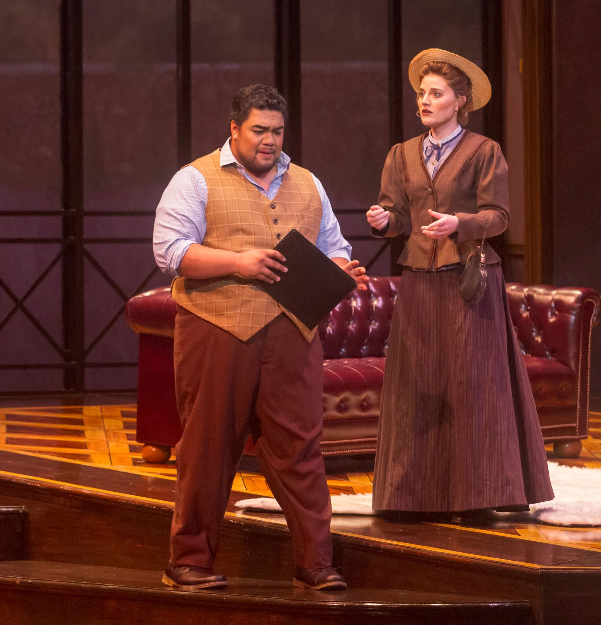 Two actors on stage during a theatrical performance. The man on the left is holding a black folder, wearing a brown vest and pants with a light blue shirt. The woman on the right is dressed in a period costume with a brown skirt, blue top, and wide-brimmed hat, speaking to the man. There is a red leather couch in the background, and the stage has wood flooring and a backdrop with a window design.