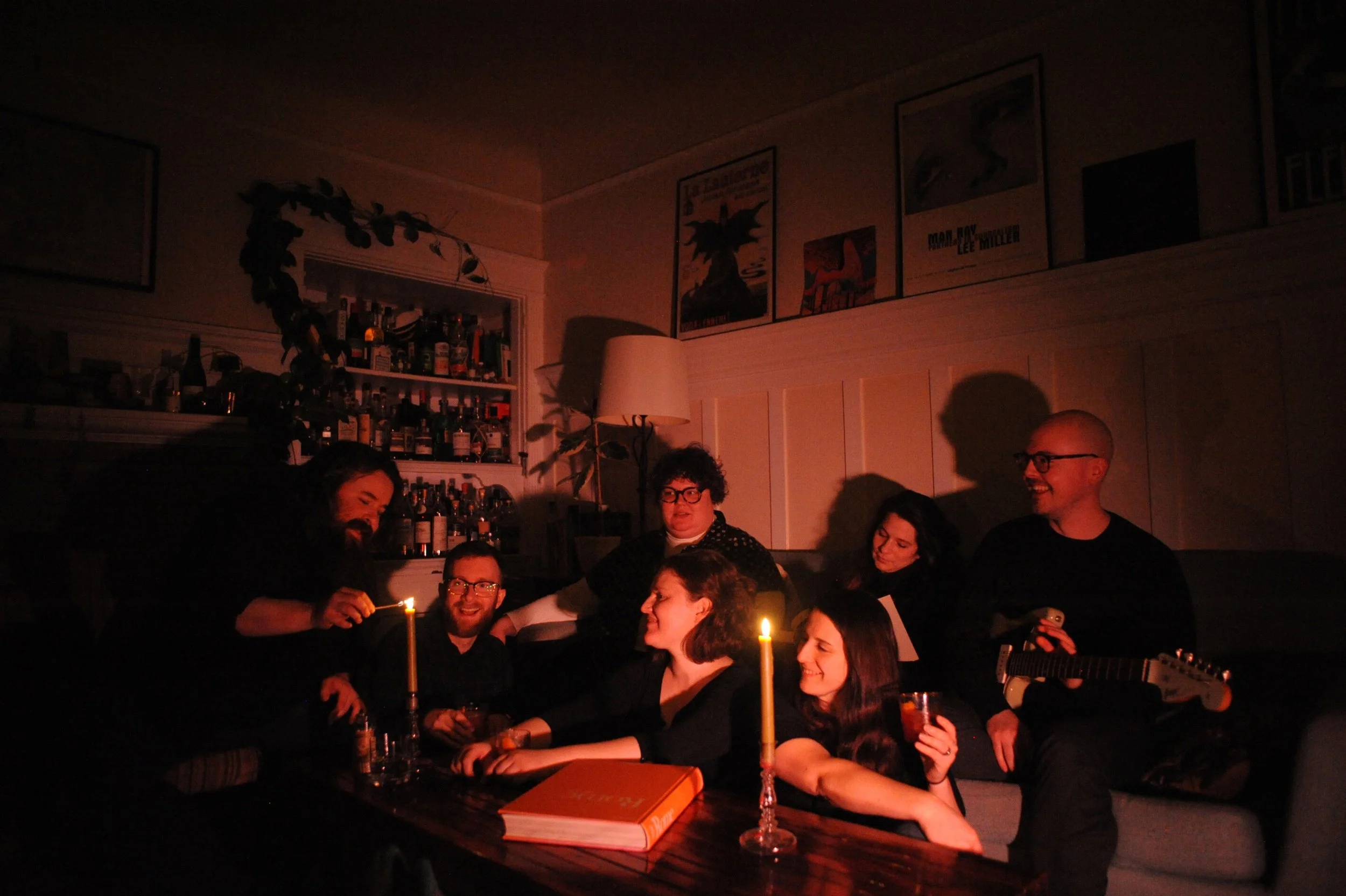 A group of seven people gathered in a cozy living room, illuminated by candlelight, enjoying drinks and conversation. One person is playing guitar, and the room features a bar with bottles on shelves, framed artwork on the walls, and a tall lamp. The atmosphere is warm and relaxed.