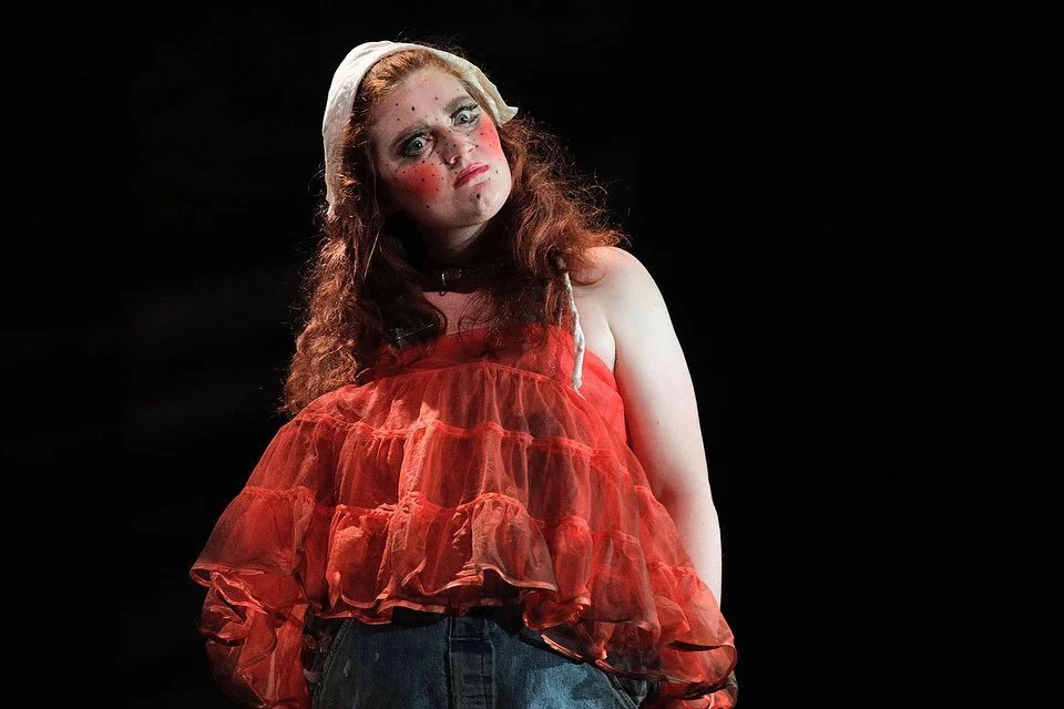 A young woman with red hair, freckles, and makeup, wearing a white headband, an orange ruffled top, and dark pants, stands against a black background.